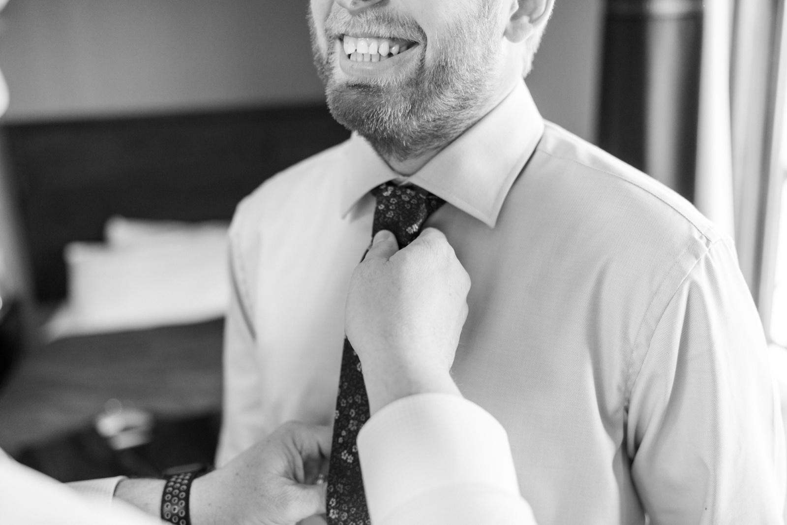 Black-and-white close-up of the groom laughing while a groomsman's hands adjust his patterned tie in soft window light — Tim Larsen Photography, Brainerd Lakes MN