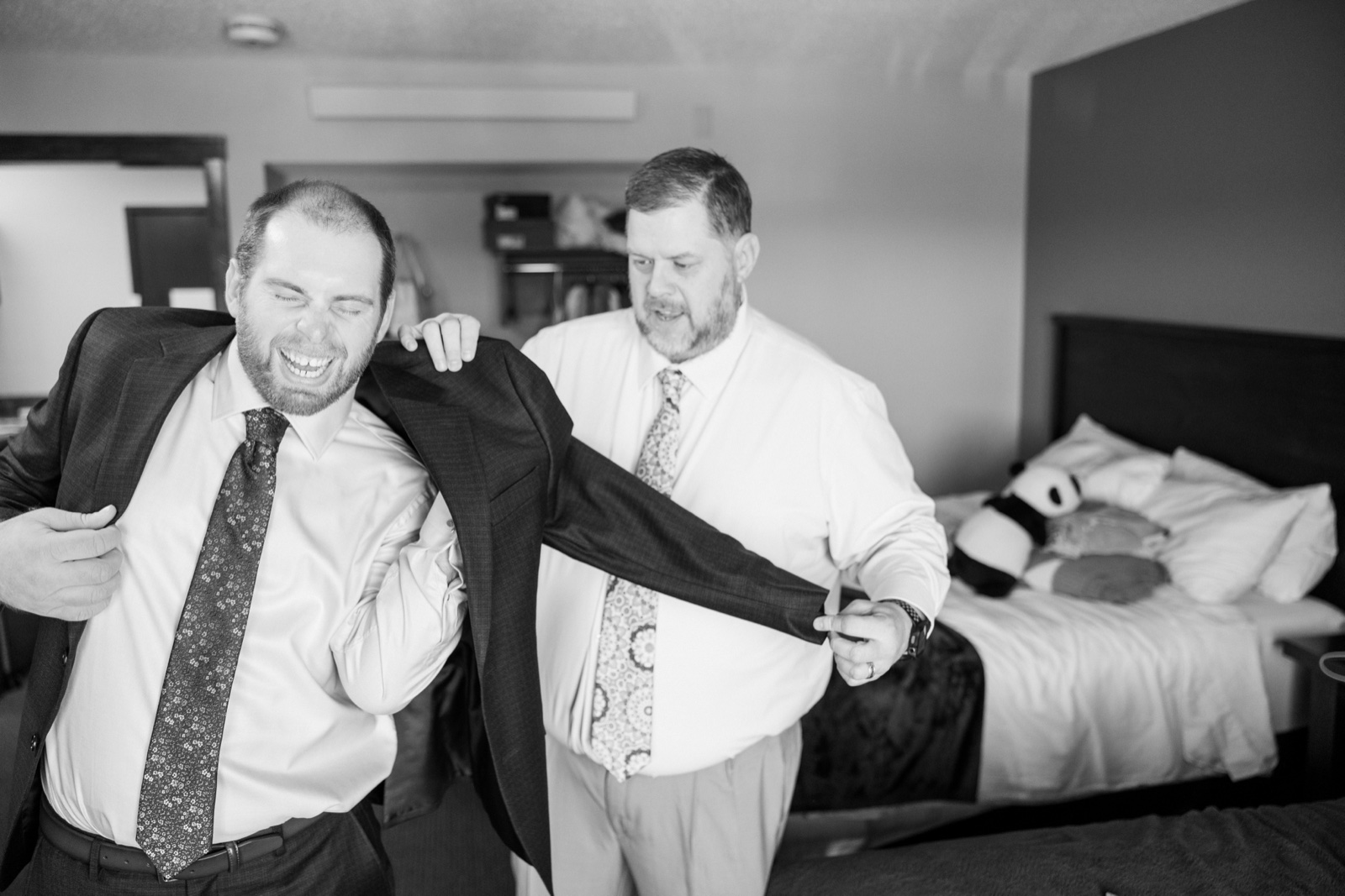 The groom laughs with his eyes shut as a groomsman helps slide his suit jacket onto his shoulders in a hotel room at Cragun's — Tim Larsen Photography, Brainerd Lakes MN