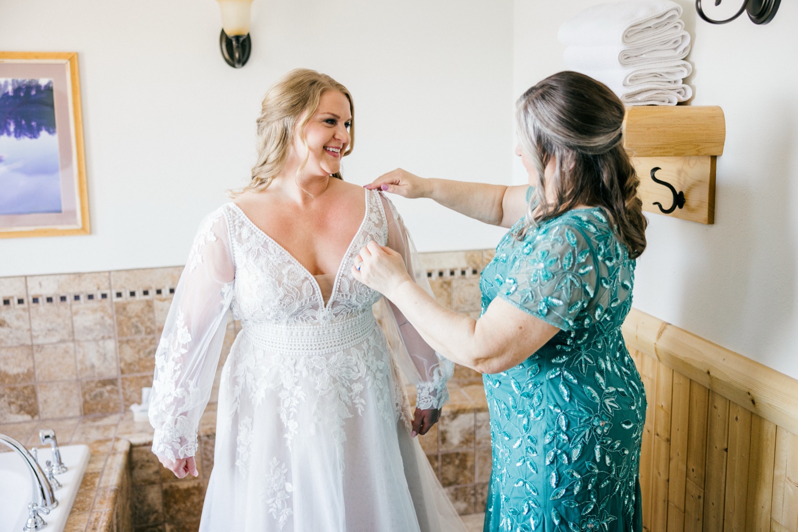 The bride smiles brightly as her mother in a teal beaded gown helps fasten the back of her lace wedding dress in the bridal suite — Tim Larsen Photography, Brainerd Lakes MN