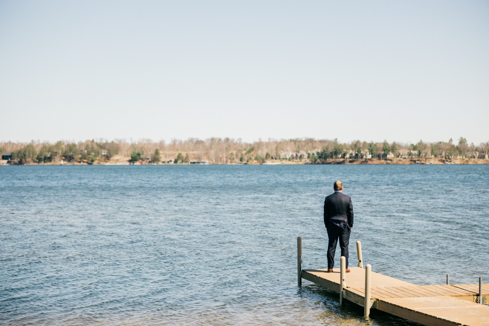 The groom in a dark suit stands alone at the end of a wooden dock looking out across Gull Lake on an overcast spring morning at Cragun's — Tim Larsen Photography, Brainerd Lakes MN