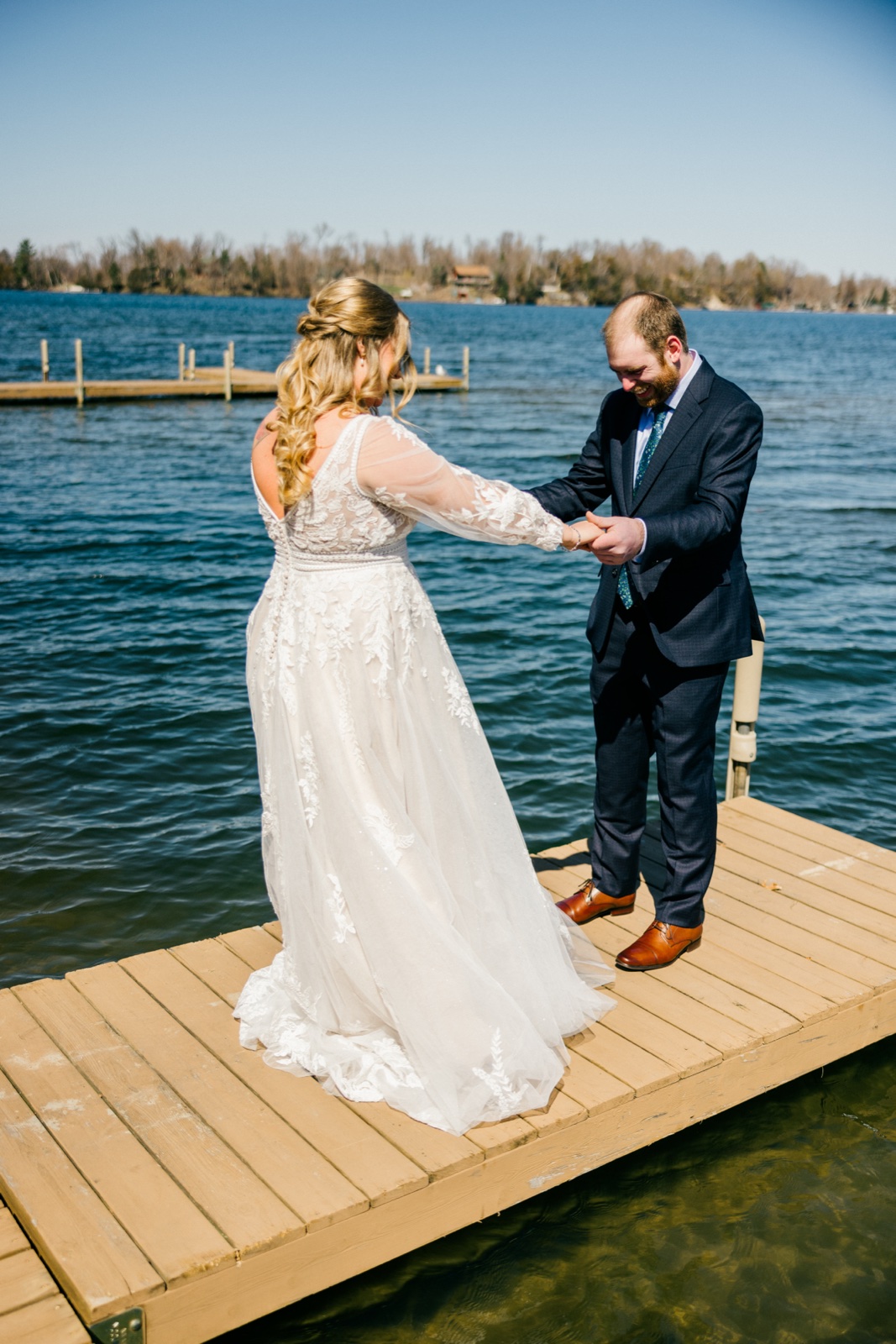 The bride and groom reach for each other's hands on the Cragun's dock during their first look, the groom smiling broadly as the bride steps toward him — Tim Larsen Photography, Brainerd Lakes MN