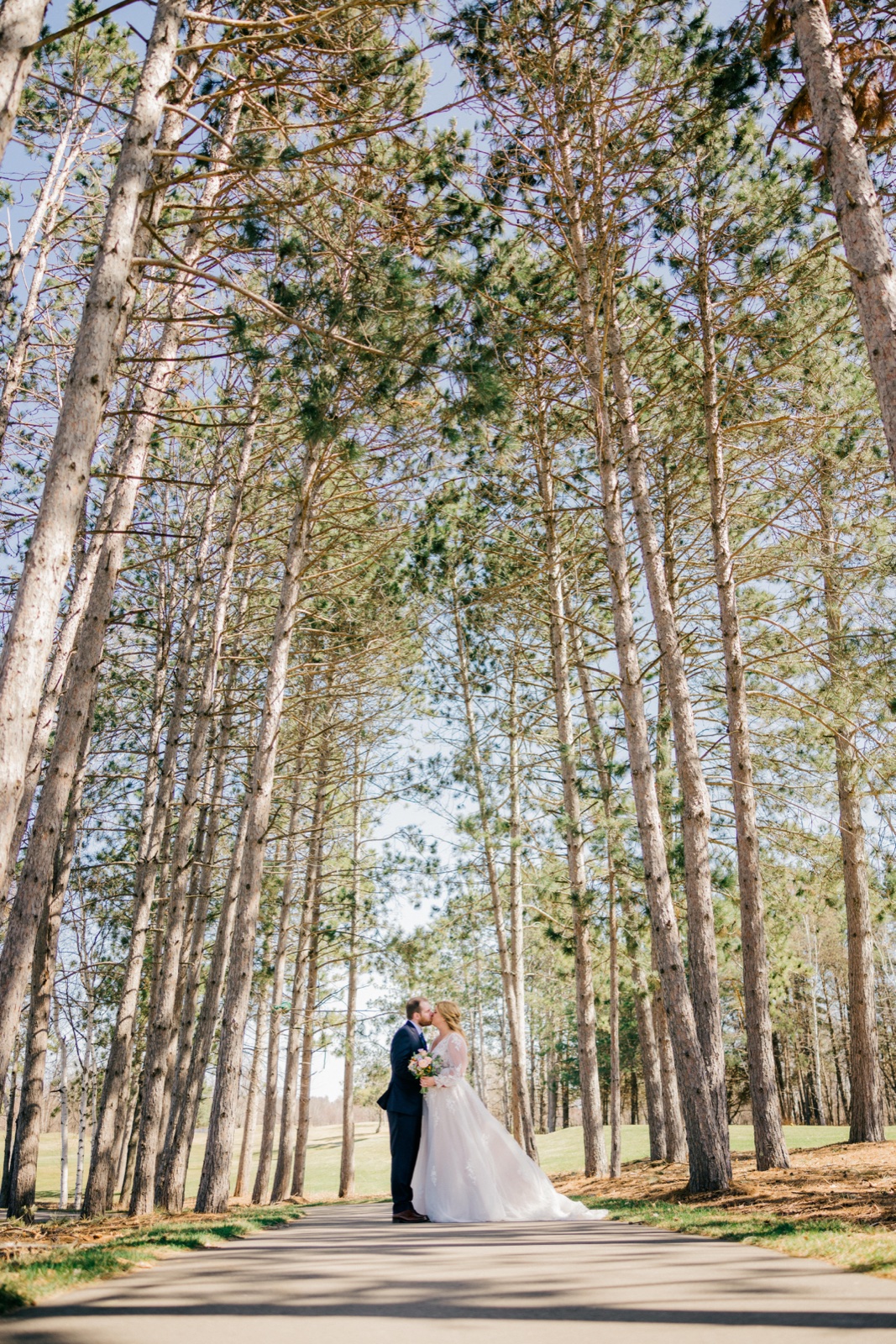 The bride and groom share a kiss while standing on a paved path beneath towering pines at Cragun's Resort — Tim Larsen Photography, Brainerd Lakes MN