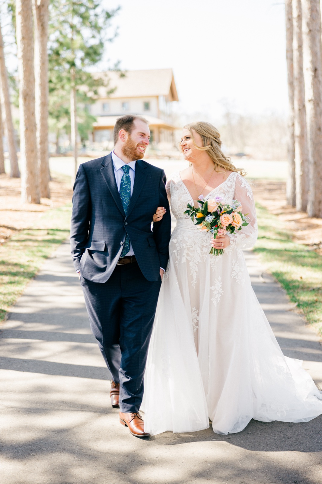 The bride and groom walk arm-in-arm down a pine-lined path at Cragun's, both laughing as they look at each other with the lodge in the background — Tim Larsen Photography, Brainerd Lakes MN