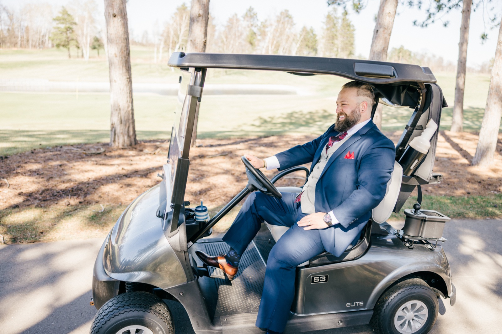The groom in a navy three-piece suit sits sideways in a golf cart on a Cragun's Resort path, laughing at someone off-camera — Tim Larsen Photography, Brainerd Lakes MN
