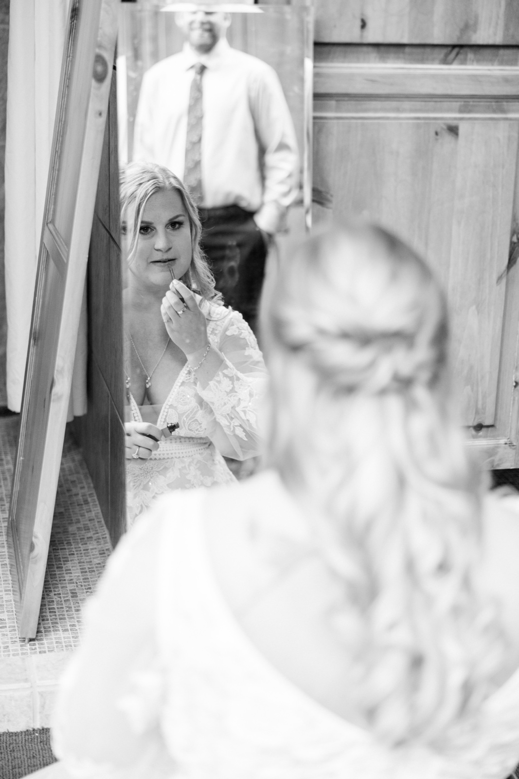 The bride applies lipstick in a mirror as the groom stands behind her in shirt and tie, the back of the bride's head soft in the foreground — Tim Larsen Photography, Brainerd Lakes MN