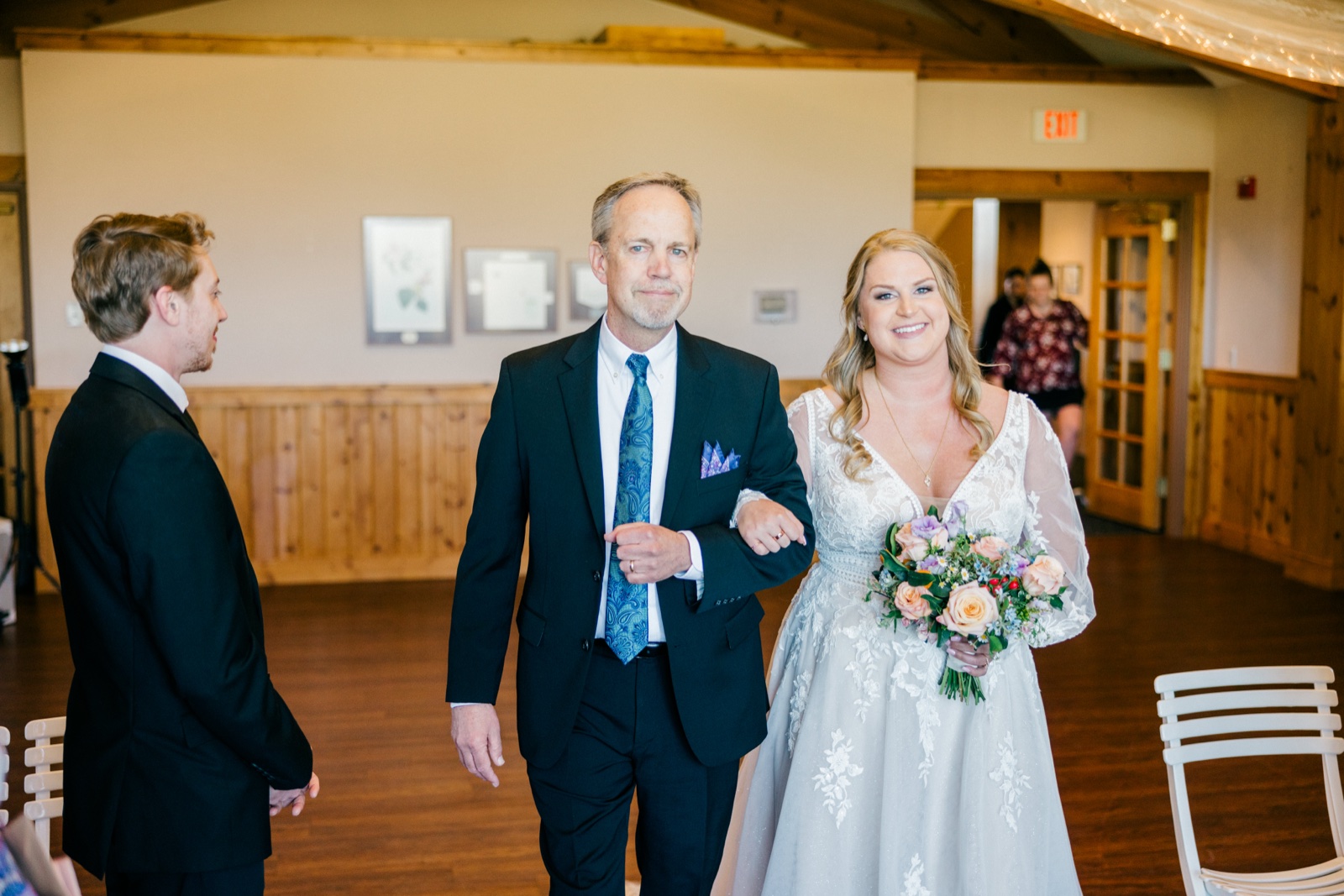 The bride walks down the aisle on her father's arm holding a pastel bouquet while the groom watches from the foreground at Cragun's — Tim Larsen Photography, Brainerd Lakes MN