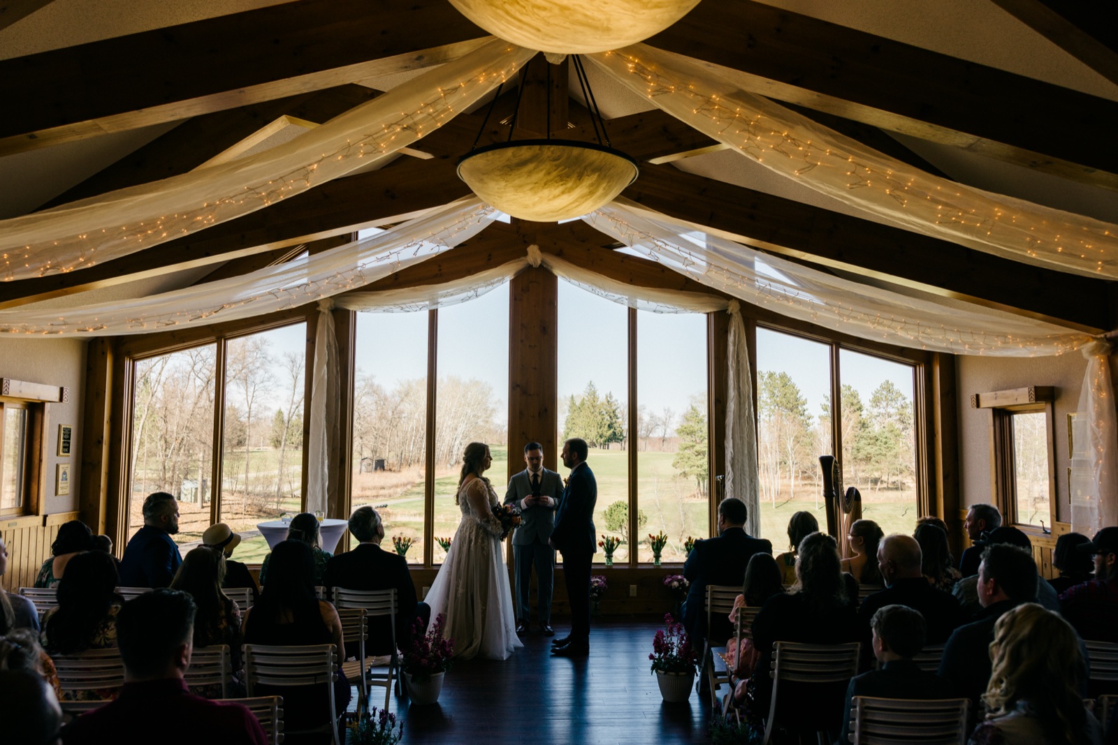 Wide frame of the bride and groom silhouetted at the front of the wood-beamed ceremony space at Cragun's, surrounded by seated guests and string lights — Tim Larsen Photography, Brainerd Lakes MN