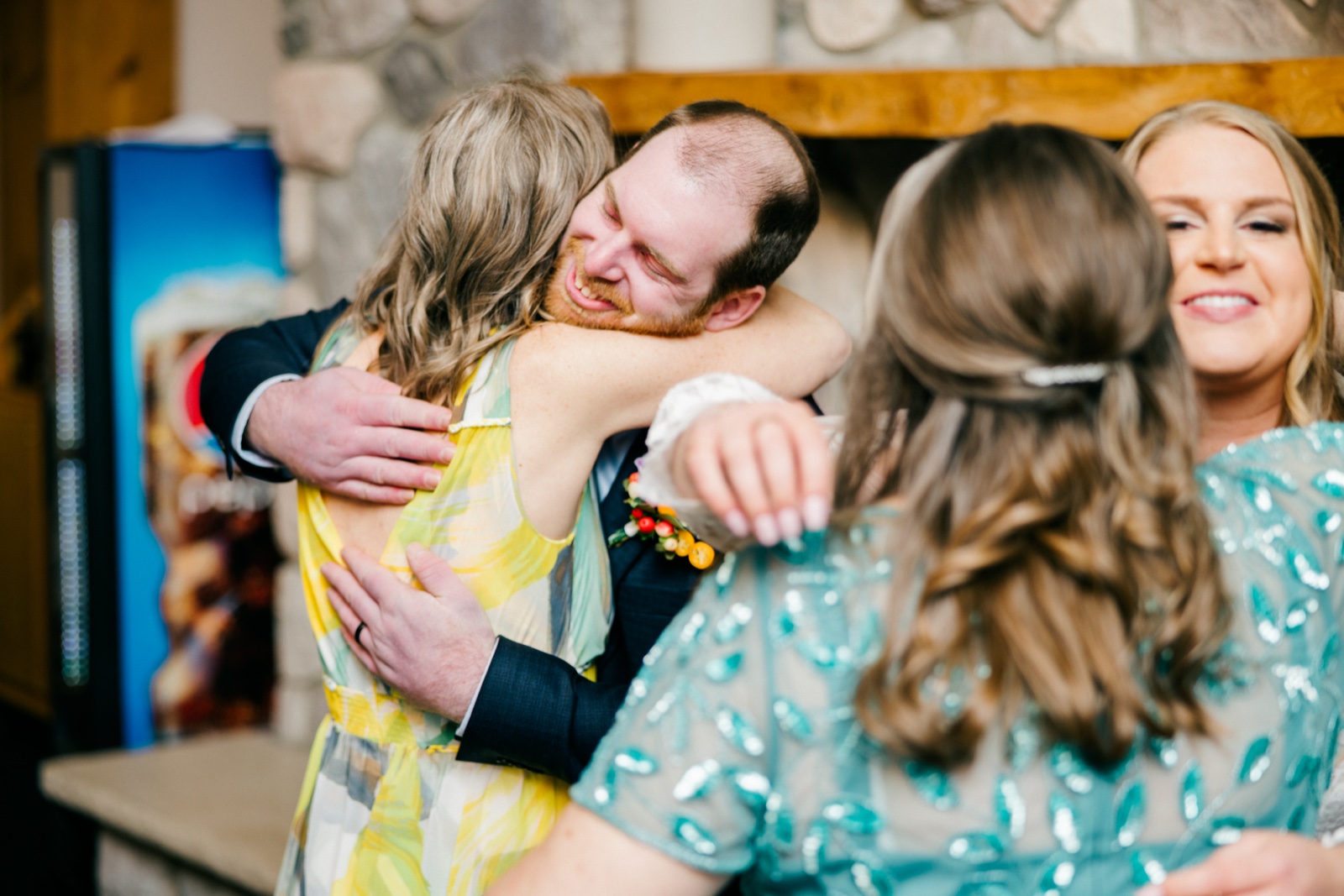 The groom closes his eyes and grins as he embraces a guest in a yellow floral dress, with a bridesmaid in teal smiling nearby at Cragun's — Tim Larsen Photography, Brainerd Lakes MN