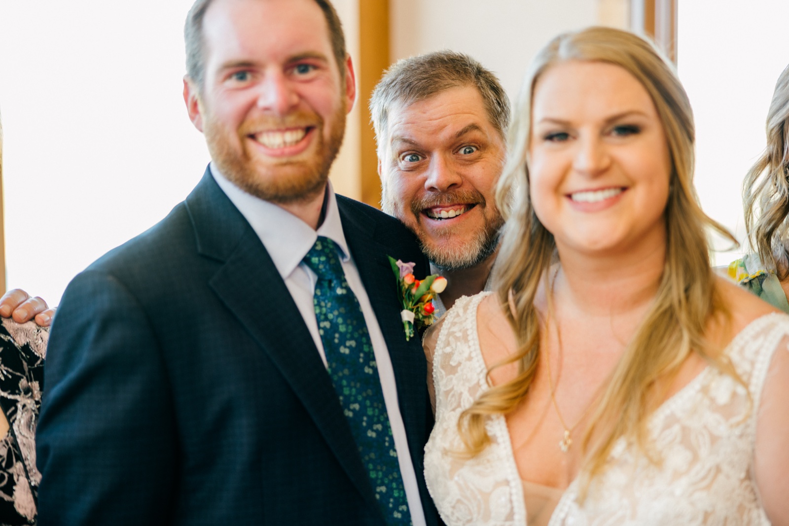The bride and groom laugh as a wide-eyed guest leans into frame between them with an exaggerated face — Tim Larsen Photography, Brainerd Lakes MN