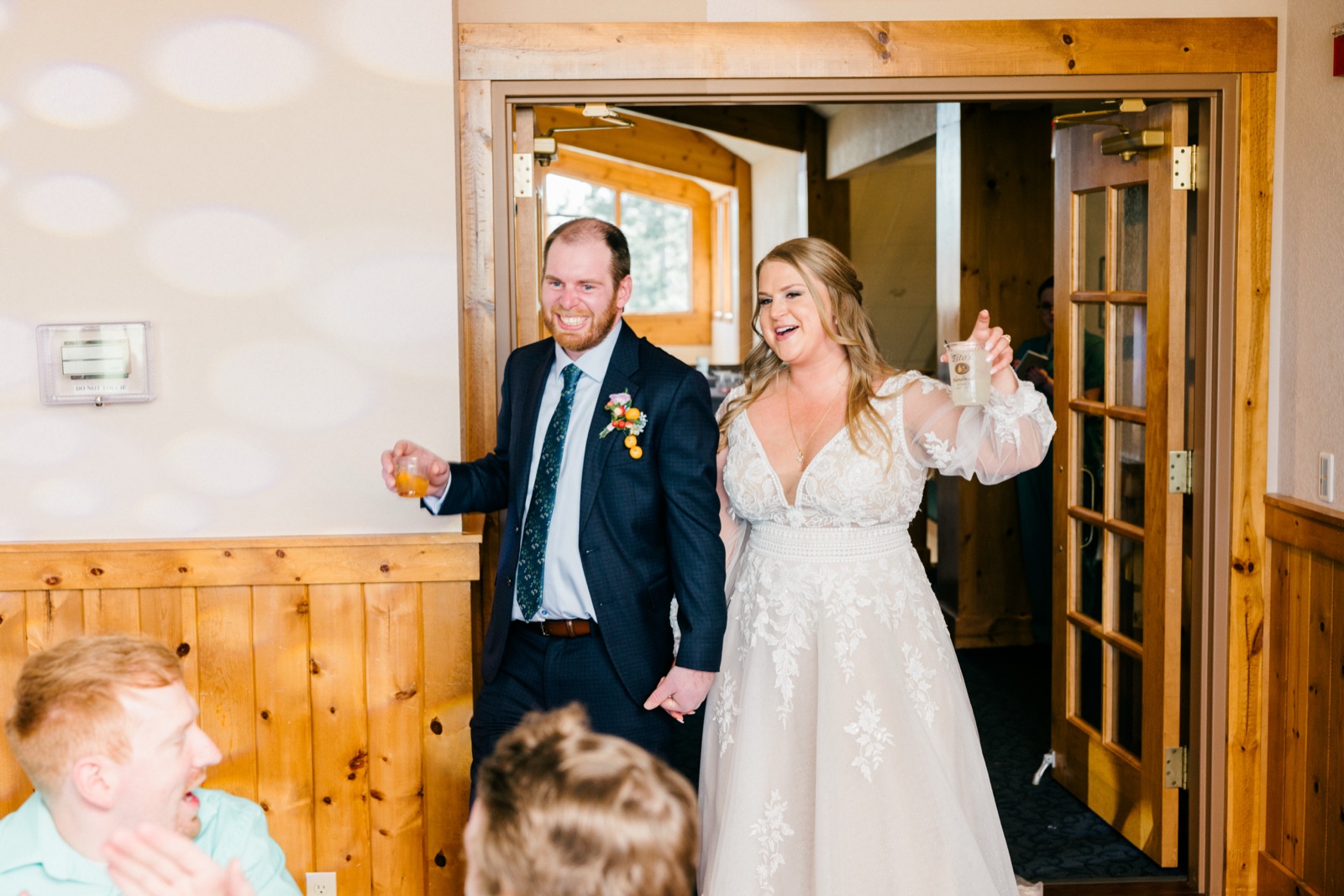 The bride and groom raise drinks and laugh as they enter their reception through a wood-framed doorway while guests cheer in the foreground — Tim Larsen Photography, Brainerd Lakes MN