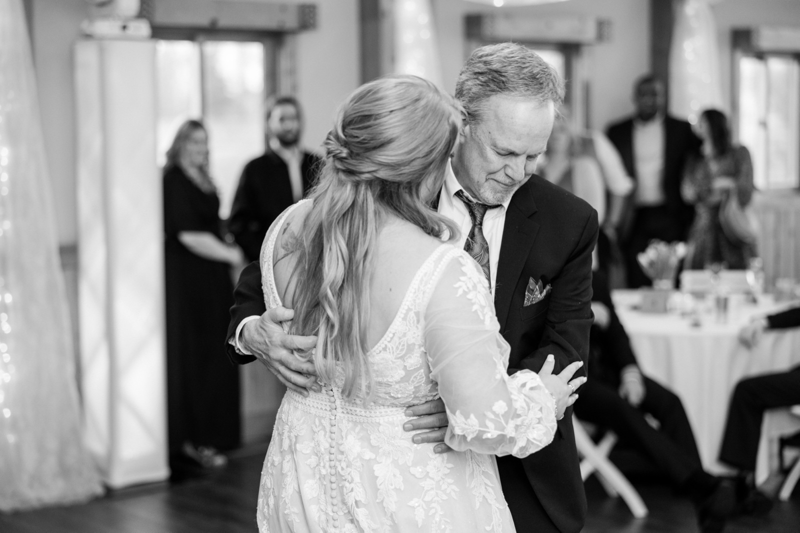 Black-and-white frame of the bride embracing her father during their dance, his eyes closed in a quiet moment as guests watch from the tables — Tim Larsen Photography, Brainerd Lakes MN