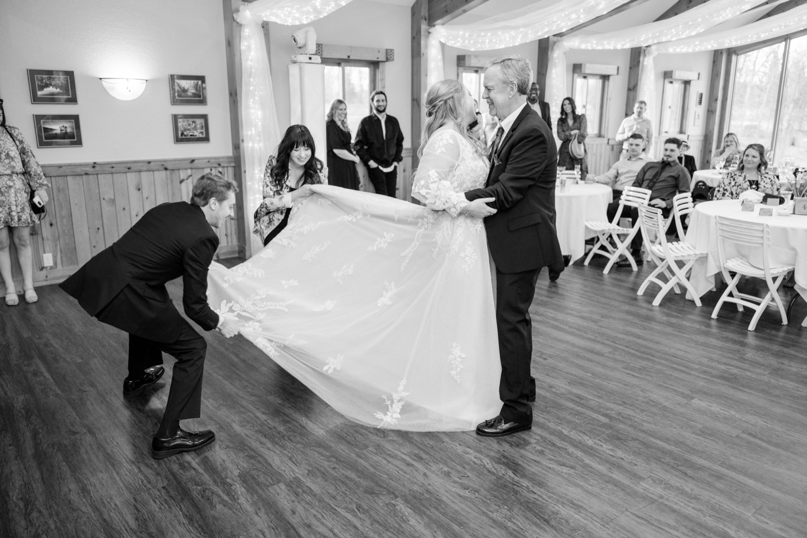 Black-and-white frame of the groom crouching to fan out the bride's lace gown as she dances with her father at Cragun's — Tim Larsen Photography, Brainerd Lakes MN