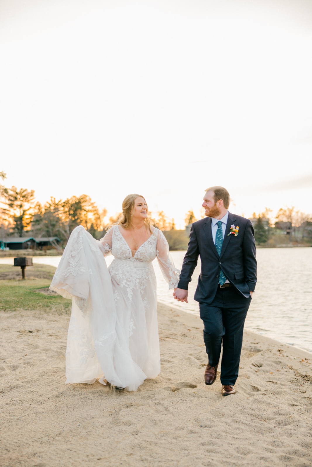 The bride lifts her gown as she walks hand-in-hand with the groom along a sandy lakeshore at sunset, both laughing — Tim Larsen Photography, Brainerd Lakes MN