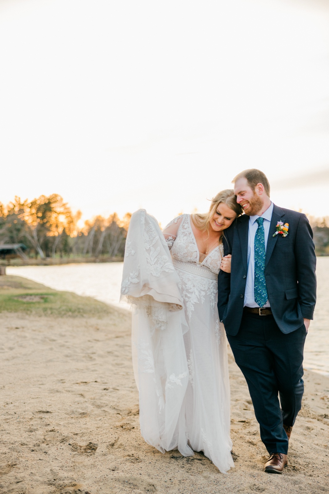 Golden-hour portrait of the bride and groom walking together along a sandy lakeshore at Cragun's Resort, the bride lifting the train of her lace gown — Tim Larsen Photography, Brainerd Lakes MN