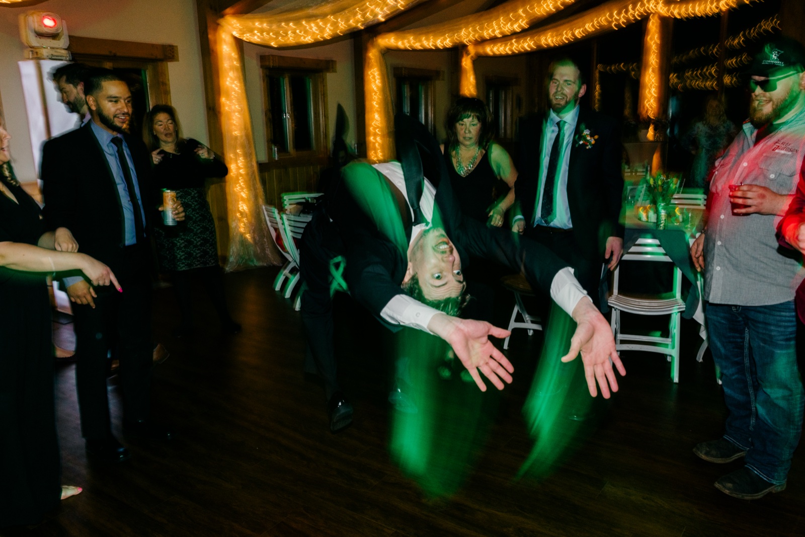 A man in a tuxedo flips upside down mid-air on the reception dance floor at Cragun's as guests watch under string lights and green stage lighting — Tim Larsen Photography, Brainerd Lakes MN