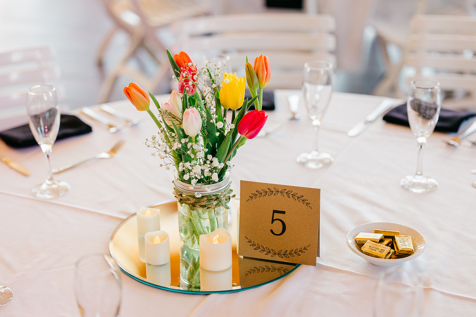 Reception table detail at Cragun's Legacy — mason jar of colorful tulips on a mirrored base with candles, kraft paper table number — Tim Larsen Photography, Brainerd Lakes MN