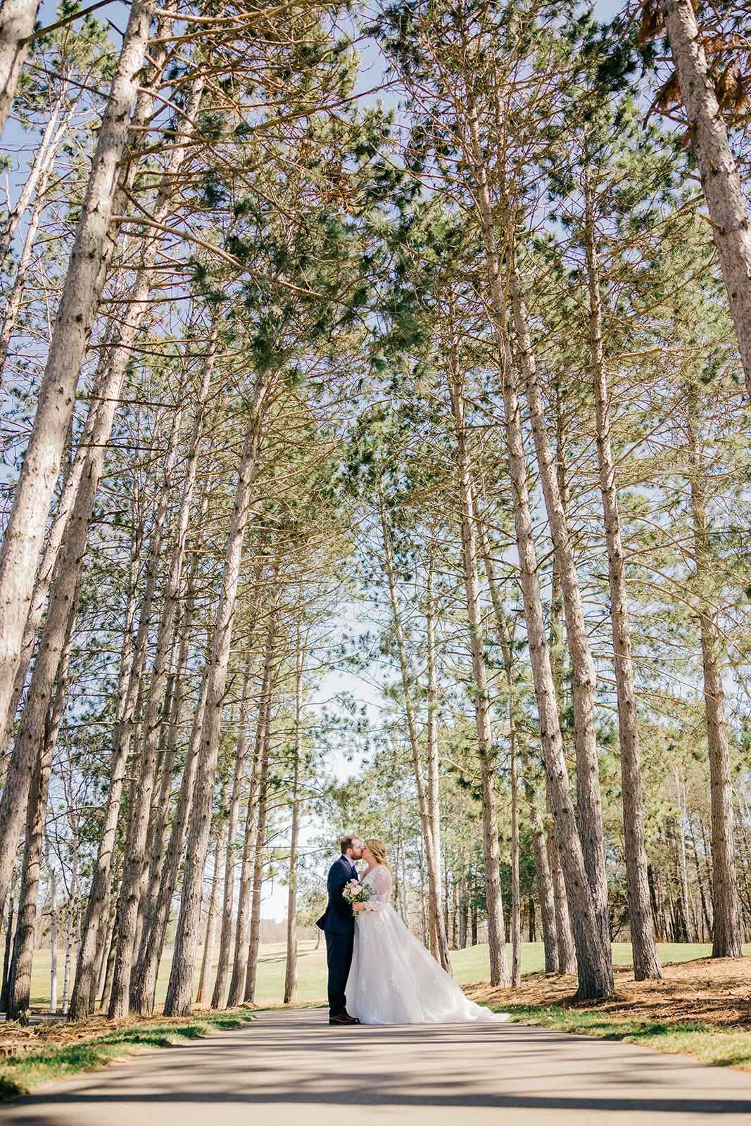Bride and groom in the pine tree corridor along the Legacy Golf Course fairway — tall pines lining both sides of the path — Tim Larsen Photography, Brainerd Lakes MN