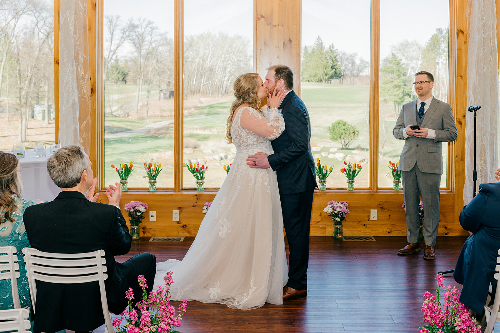 First kiss in the Audubon Room at Cragun's Legacy Golf Course, guests applauding, golf course visible through the windows — Tim Larsen Photography, Brainerd Lakes MN