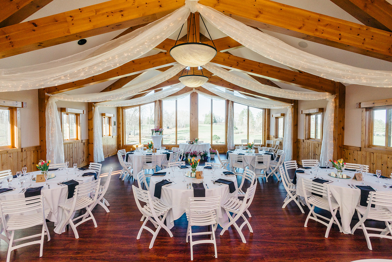 Audubon Room reception setup at Cragun's Legacy Golf Course — round tables with white linens, navy napkins, tulip centerpieces, twinkle lights on timber beams — Tim Larsen Photography, Brainerd Lakes MN