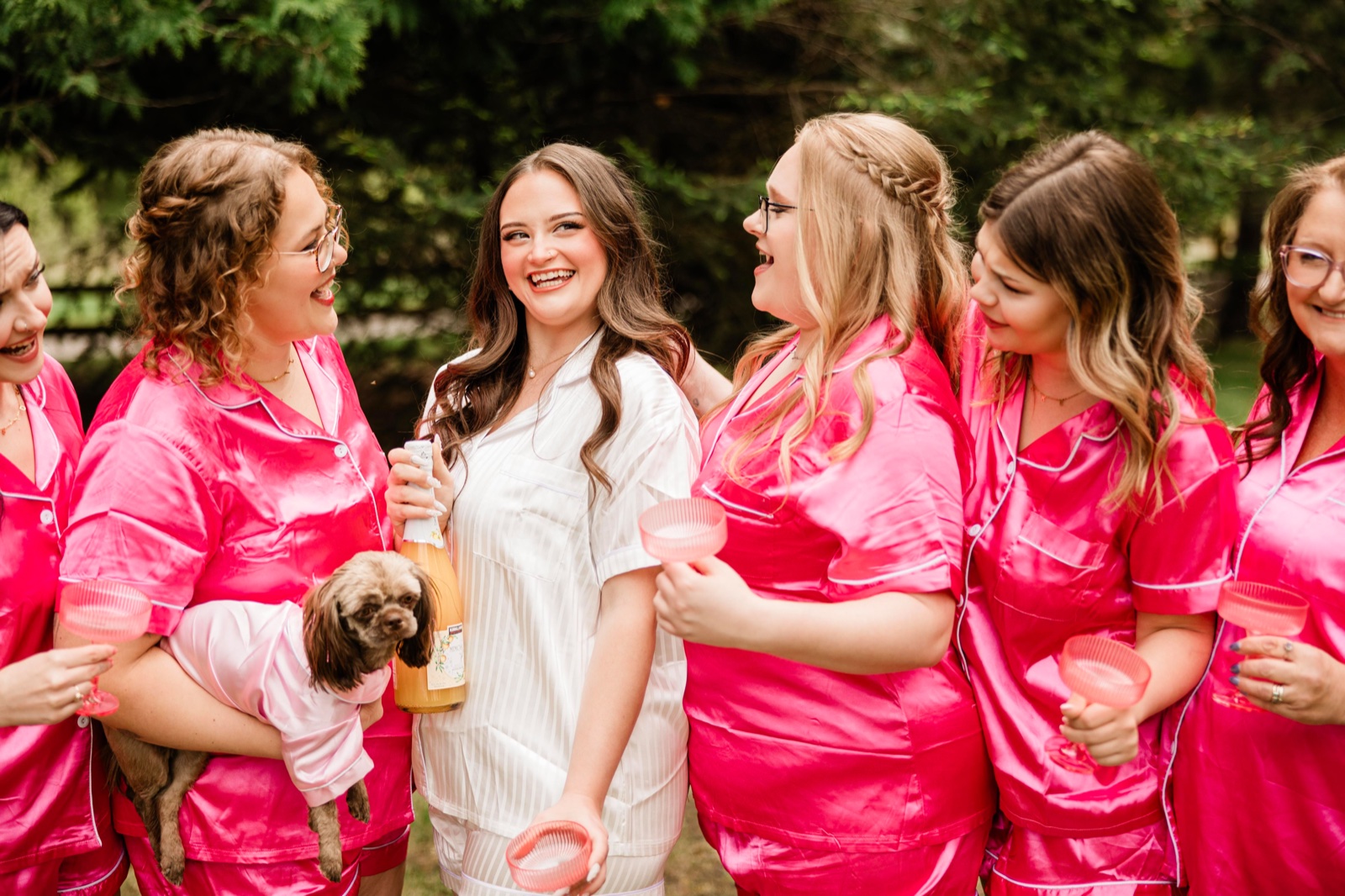 Bride laughing with bridesmaids in hot-pink satin pajamas, a small dog in someone's arms
