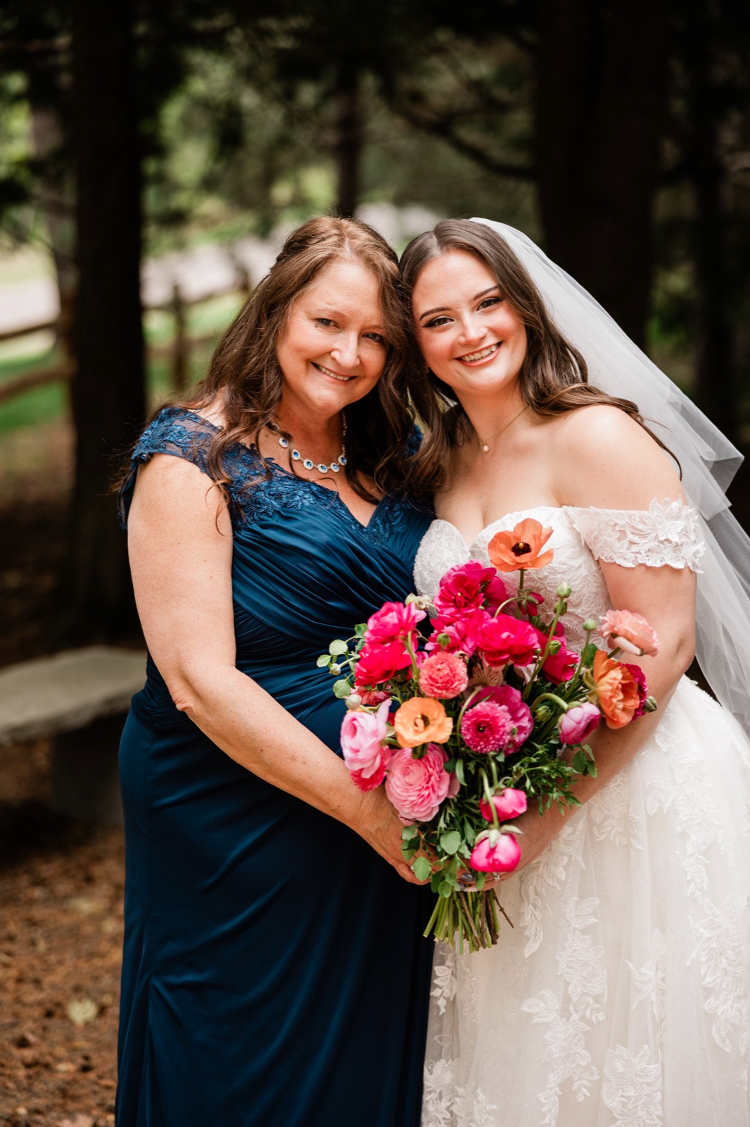 Bride cheek-to-cheek with her mother in the pines, coral and pink bouquet between them