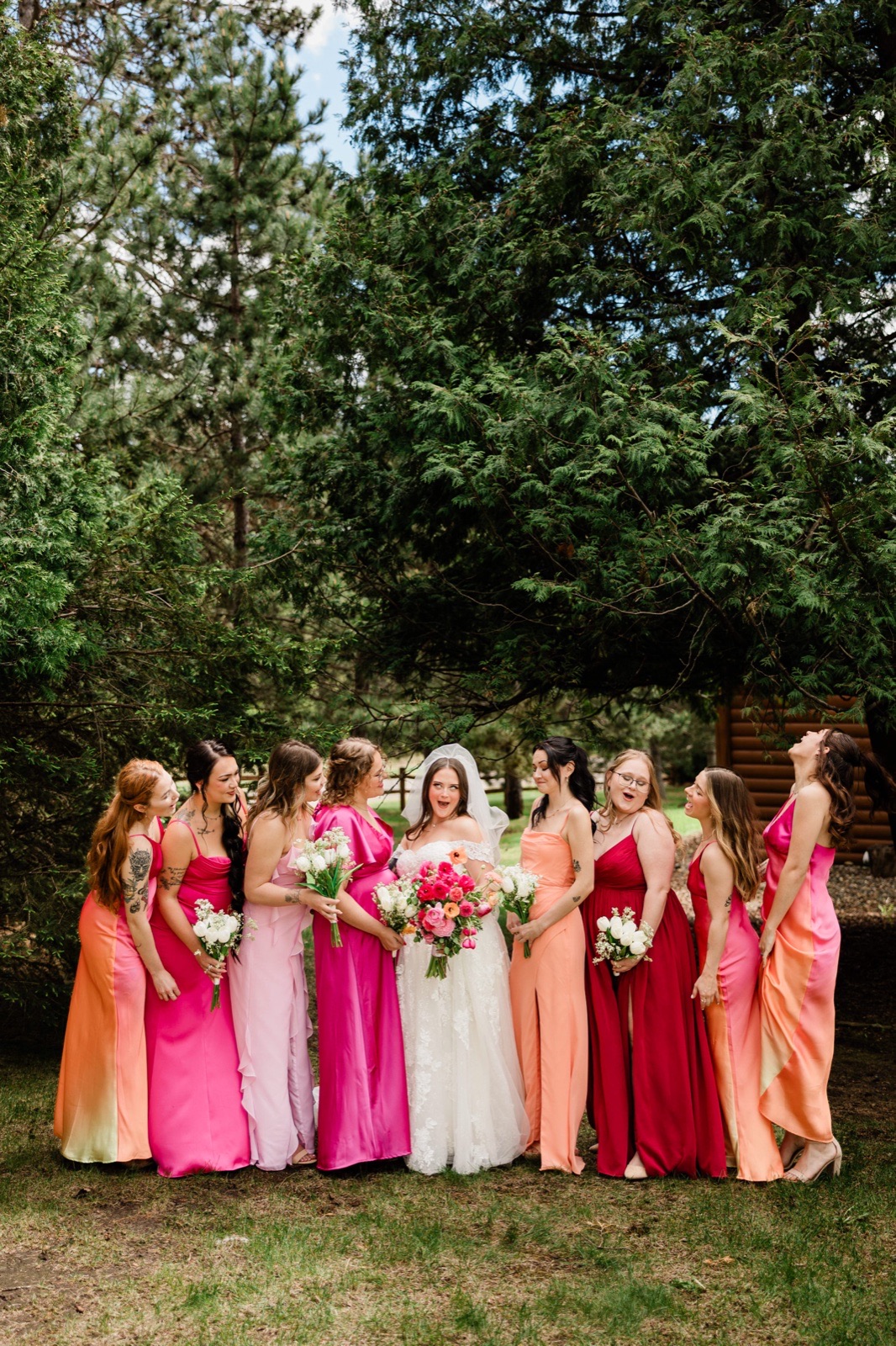 Bride and bridesmaids in pink, coral and magenta laughing under tall pines near a log cabin