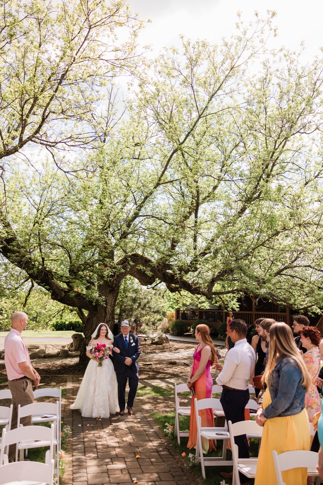 Bride walking the brick aisle with her father under an enormous old tree at Pine Peaks Event Center