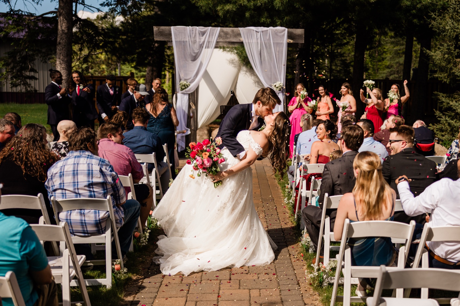 The kiss mid-aisle as guests shower the couple with flower petals beneath the draped wooden arch