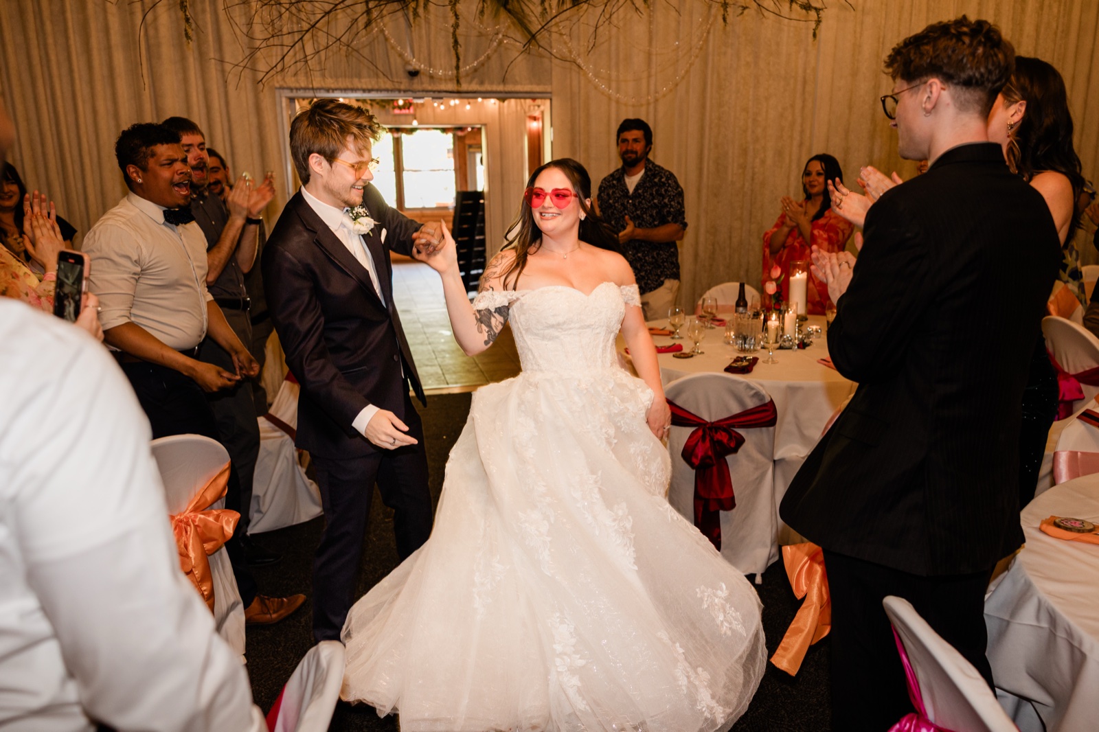 Bride in pink heart-shaped sunglasses twirling as the groom and guests cheer on the dance floor