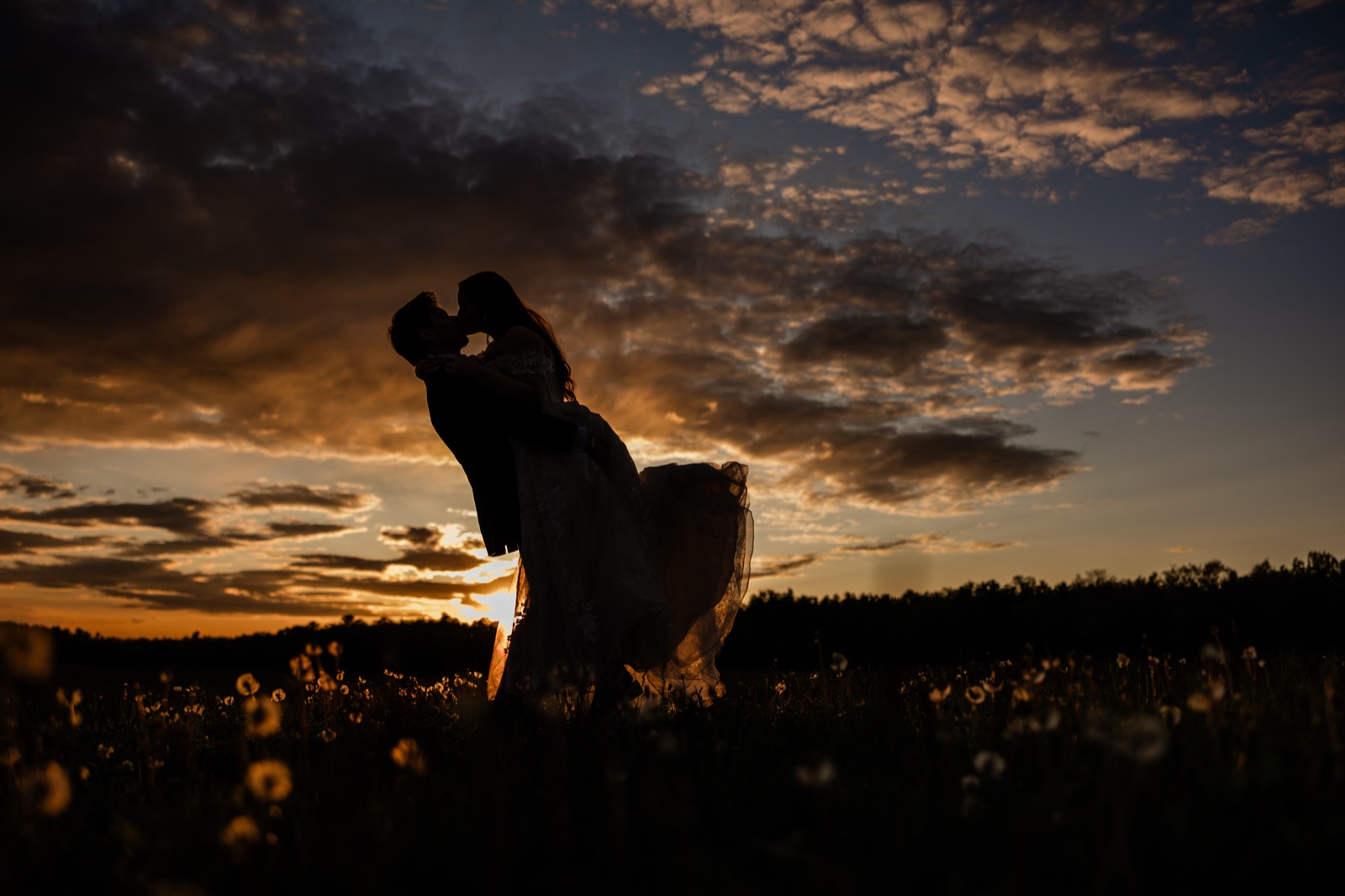Silhouettes of the couple kissing against a blazing orange sunset sky over the Pine Peaks meadow
