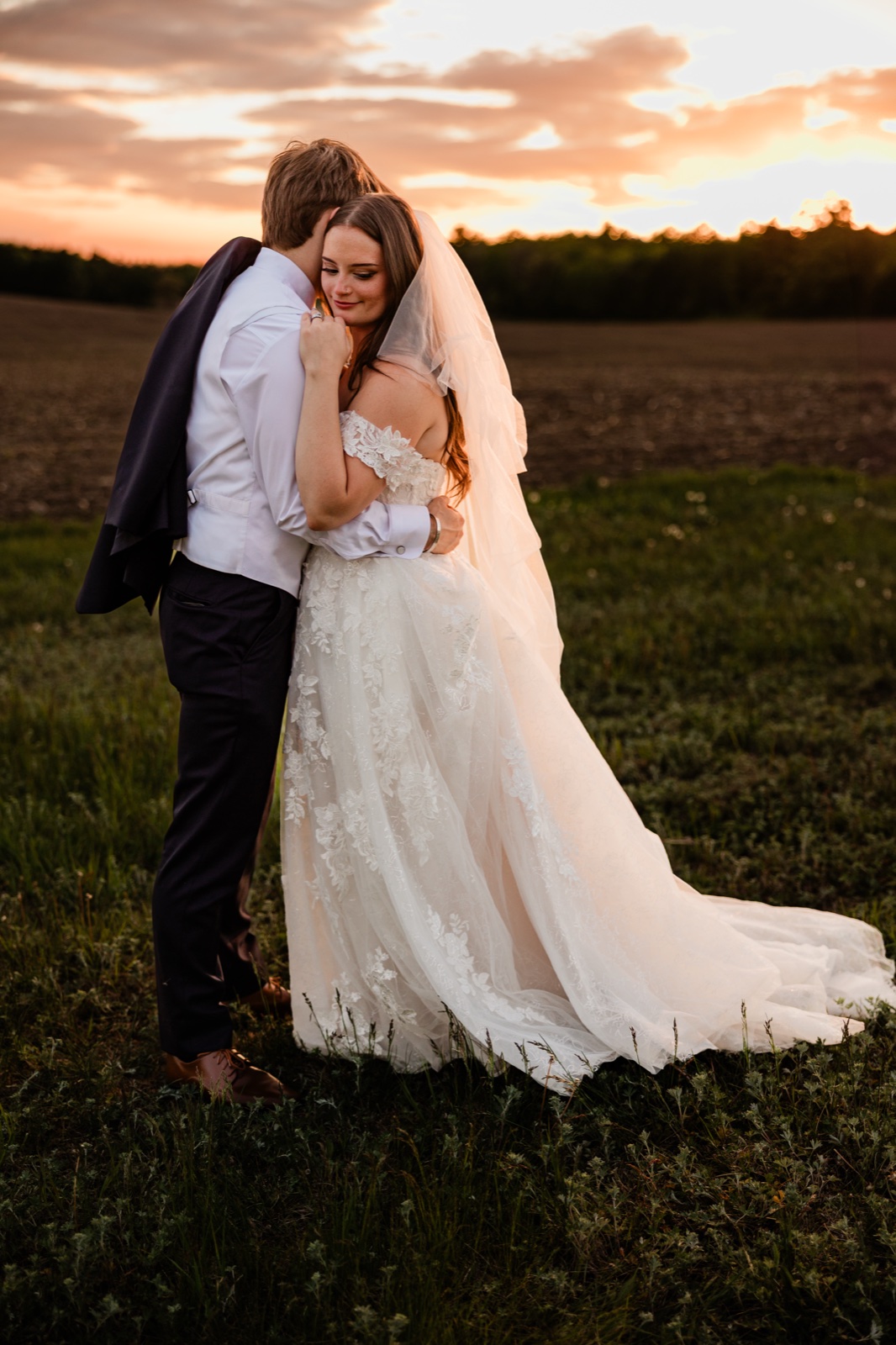 Bride resting her head on the groom's chest in an open field beneath a vivid sunset sky