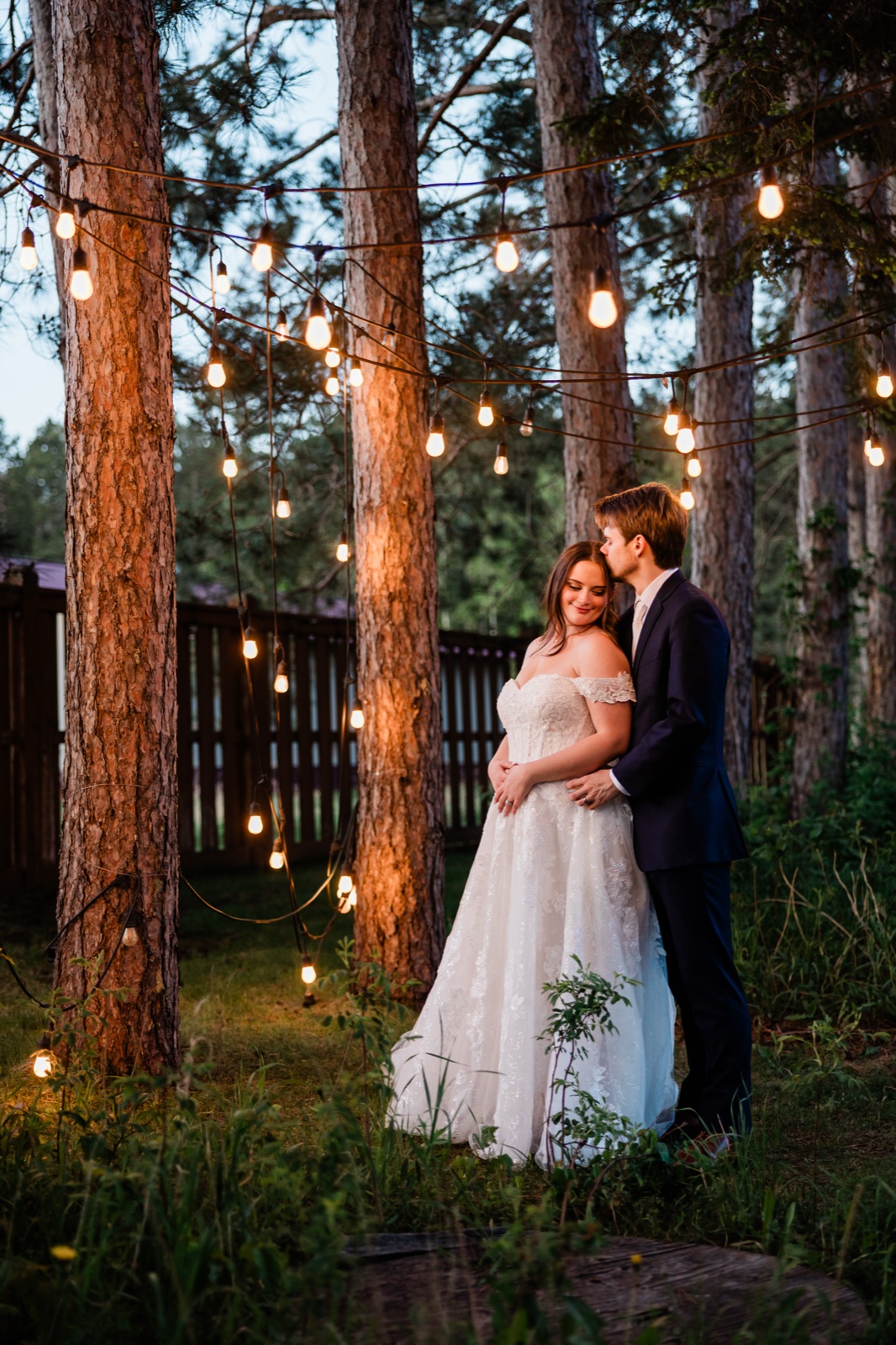 Groom kissing the bride's forehead under the Edison-bulb canopy at Pine Peaks Event Center