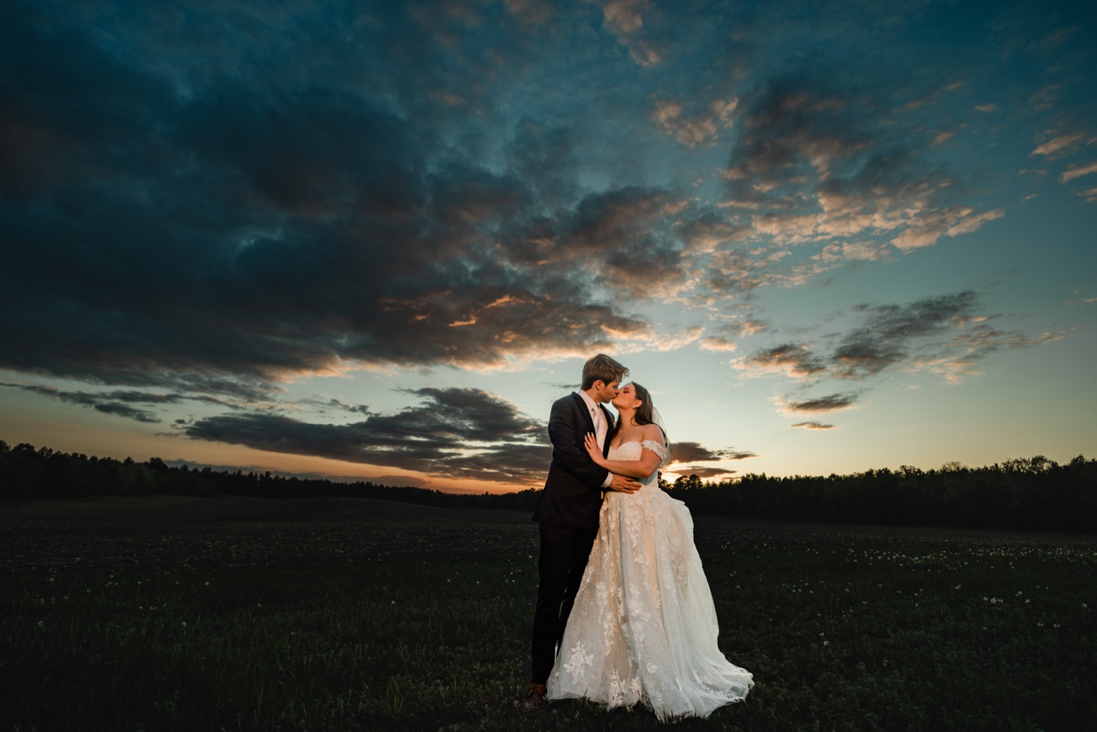 The couple small against an enormous sunset sky in an open field at Pine Peaks