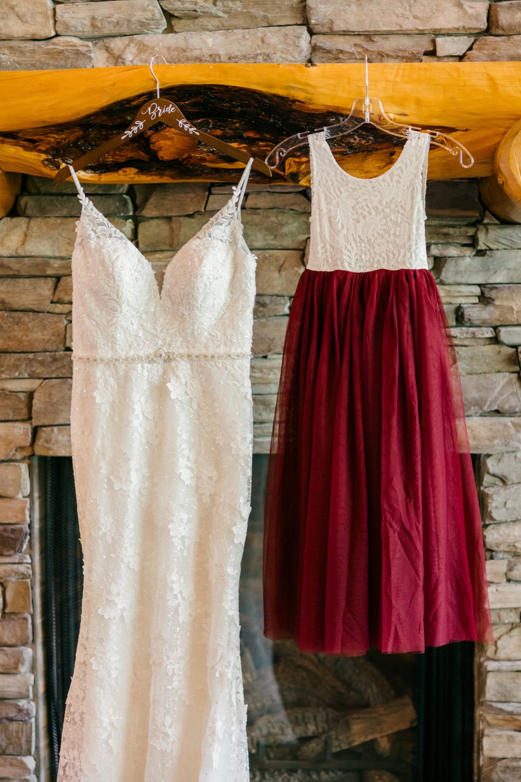 A white lace bridal gown and a burgundy tulle flower girl dress hang side-by-side from a live-edge wooden mantel above a stone fireplace — Tim Larsen Photography, Brainerd Lakes MN
