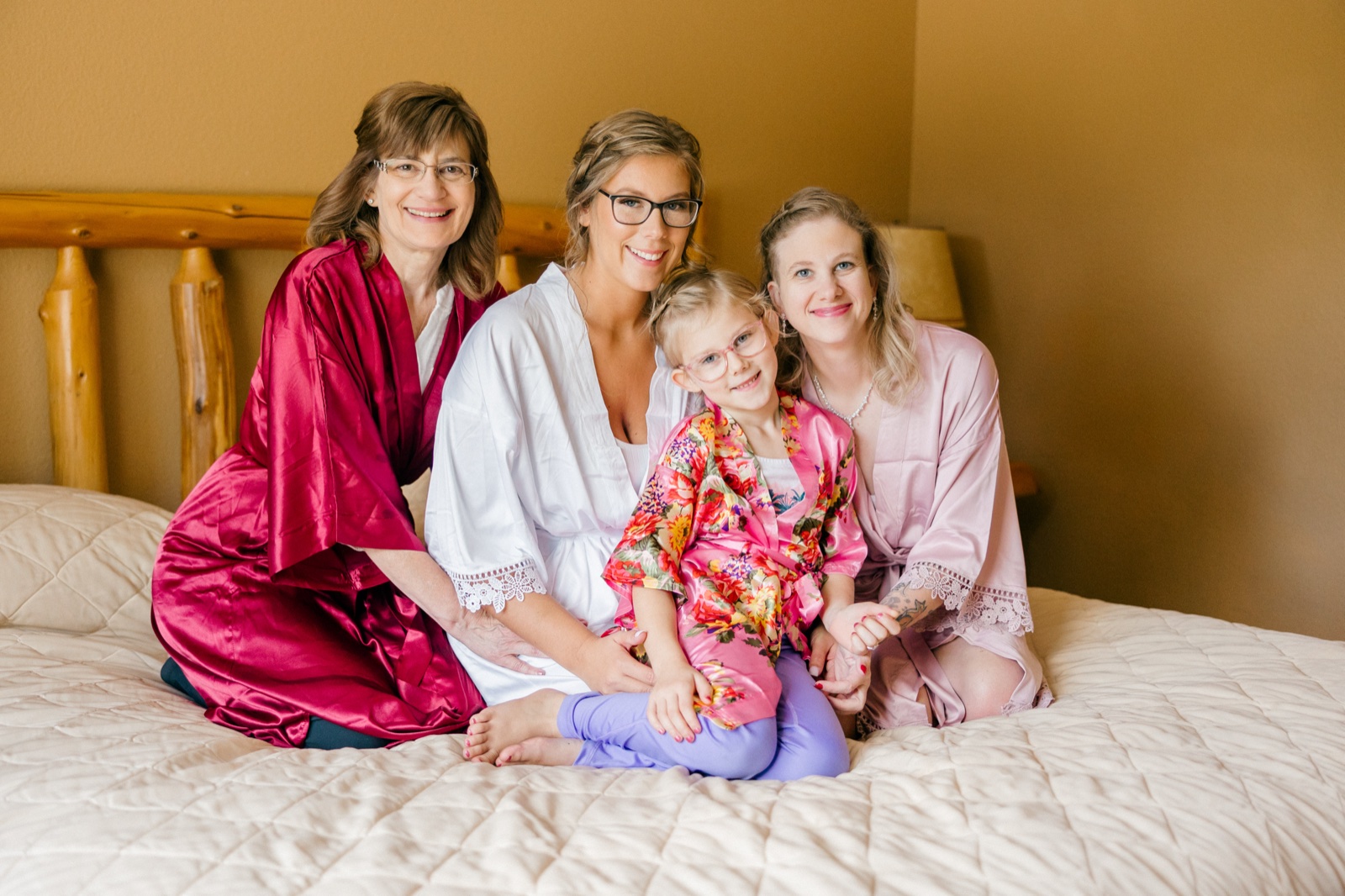 Kim sits on a bed in a white robe surrounded by her mother, sister, and a young flower girl, all in coordinating robes and smiling at the camera — Tim Larsen Photography, Brainerd Lakes MN