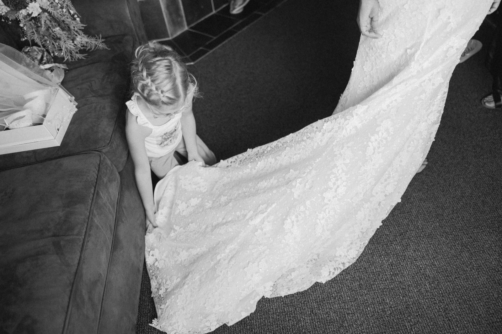 A young flower girl crouches on a couch to examine the long lace train of Kim's dress as it sweeps across the floor — Tim Larsen Photography, Brainerd Lakes MN