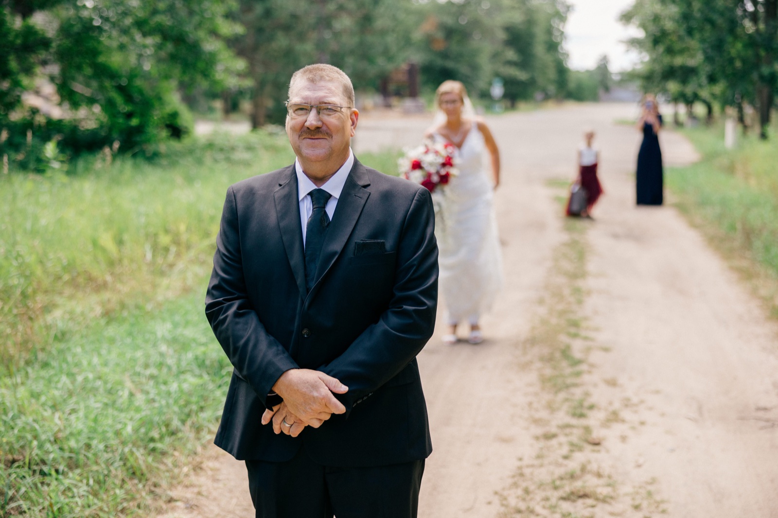 Kim's father in a dark suit waits on a dirt road as the bride approaches from behind, holding her bouquet — Tim Larsen Photography, Brainerd Lakes MN