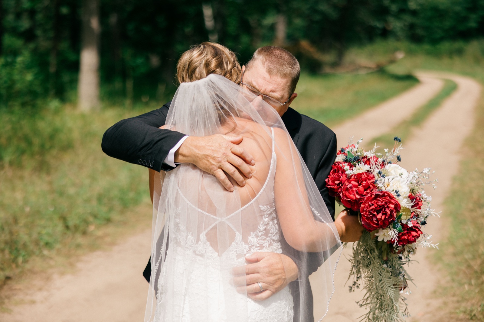 Kim embraces her father on a wooded dirt road, her veil draped across her back as she holds a red and white bouquet to the side — Tim Larsen Photography, Brainerd Lakes MN