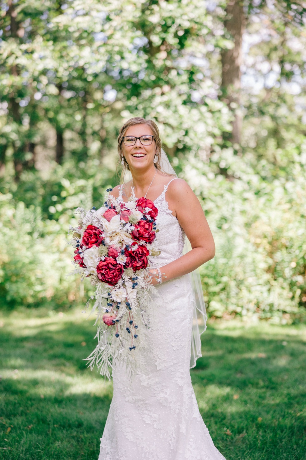 Kim laughs while holding a large cascading red and white bouquet, her veil draped over one shoulder against a wooded backdrop — Tim Larsen Photography, Brainerd Lakes MN