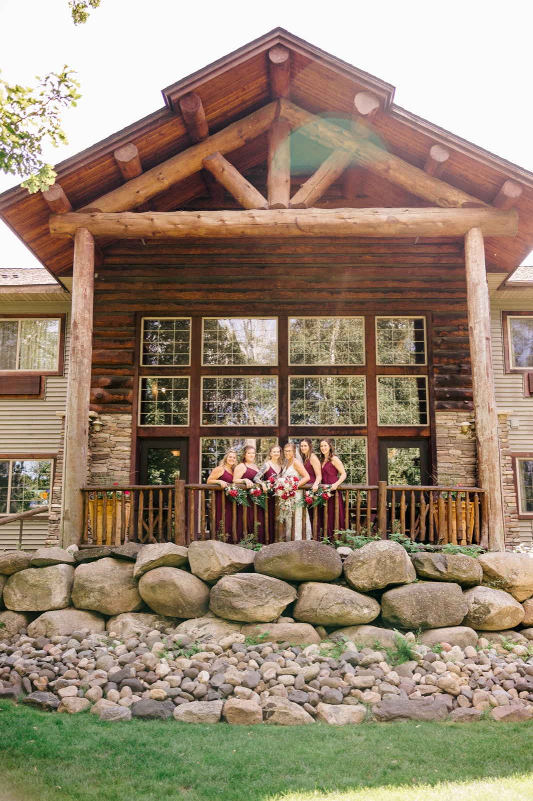 Kim and her bridesmaids in burgundy gowns stand on the wooden porch of Whitefish Lodge beneath a tall log-truss gable — Tim Larsen Photography, Brainerd Lakes MN