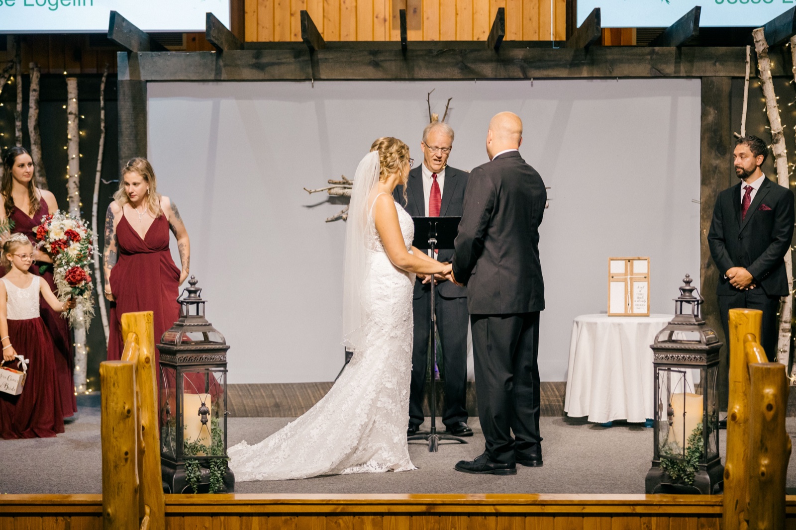 Kim and Jesse hold hands facing the officiant on a wooden ceremony stage flanked by birch trunks and large lanterns, with bridesmaids and groomsmen watching — Tim Larsen Photography, Brainerd Lakes MN