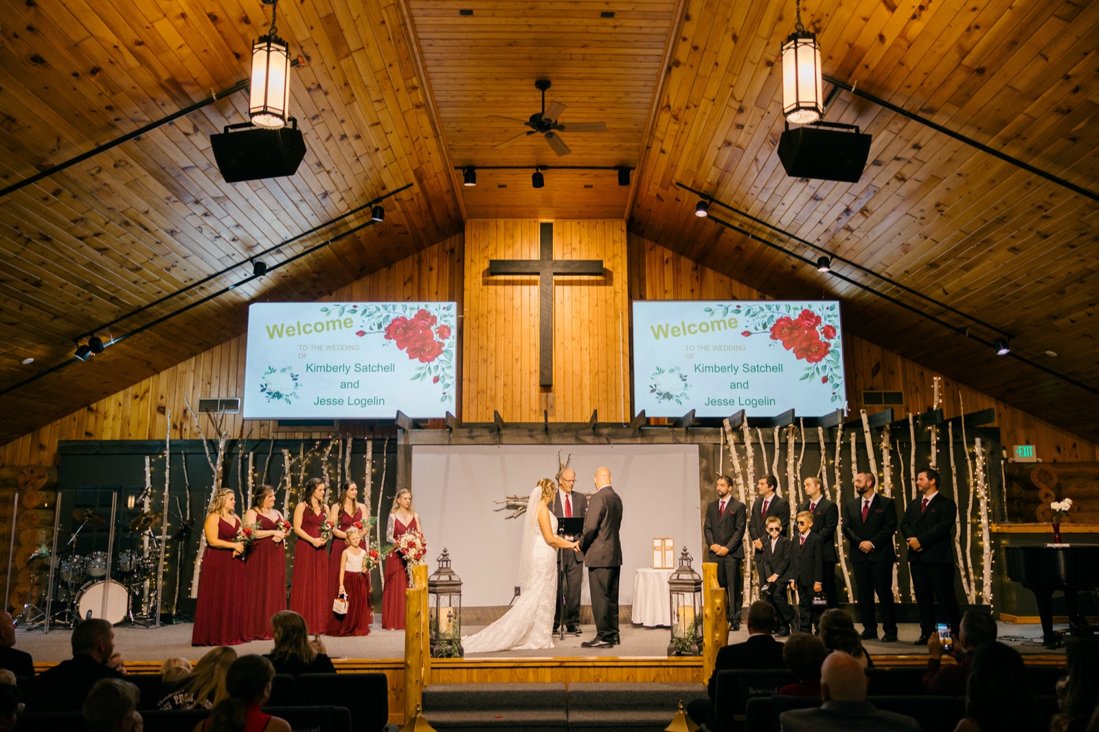 A wide symmetrical view of the ceremony in a wood-paneled chapel with the couple at center facing the officiant and the bridal party arranged on either side beneath a tall cross — Tim Larsen Photography, Brainerd Lakes MN