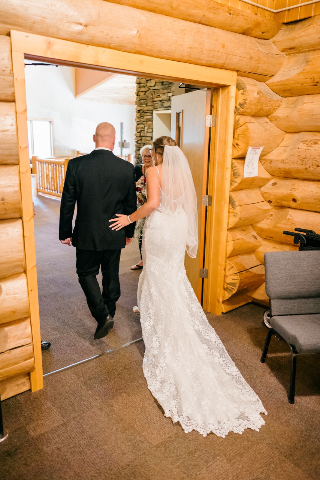 The bride walks toward the ceremony through a log-framed doorway, her lace train spread across the carpet, as the groom waits ahead — Tim Larsen Photography, Brainerd Lakes MN