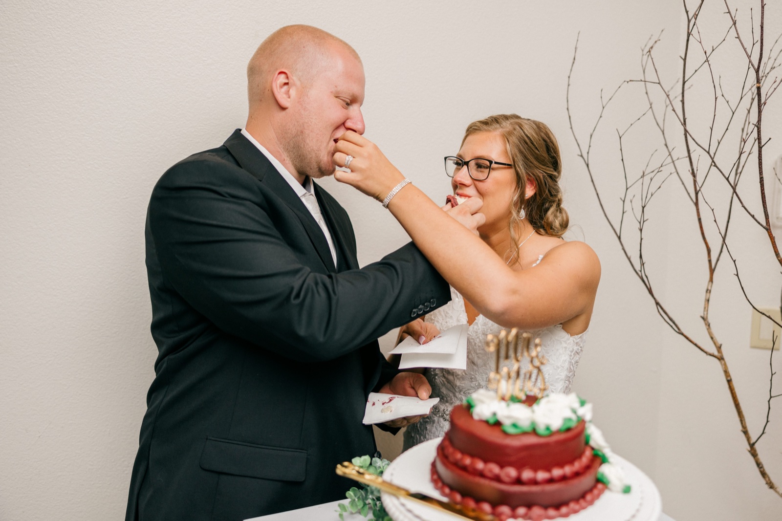 The bride laughs as she pushes a piece of red cake into the groom's face during the cake cutting at their reception — Tim Larsen Photography, Brainerd Lakes MN