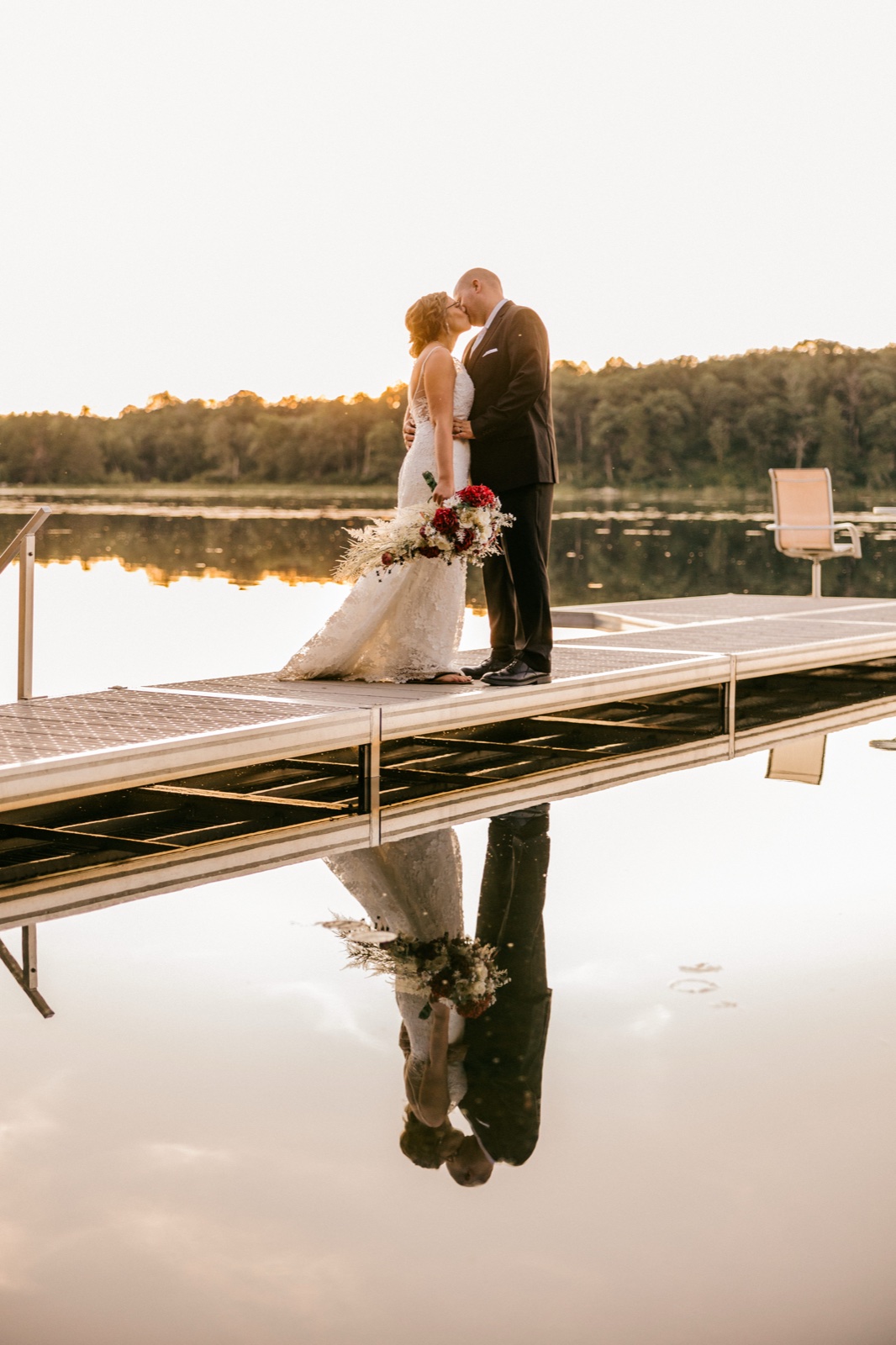 Kim and Jesse share a kiss at the end of a dock at golden hour, their figures mirrored in the glassy surface of the lake below — Tim Larsen Photography, Brainerd Lakes MN