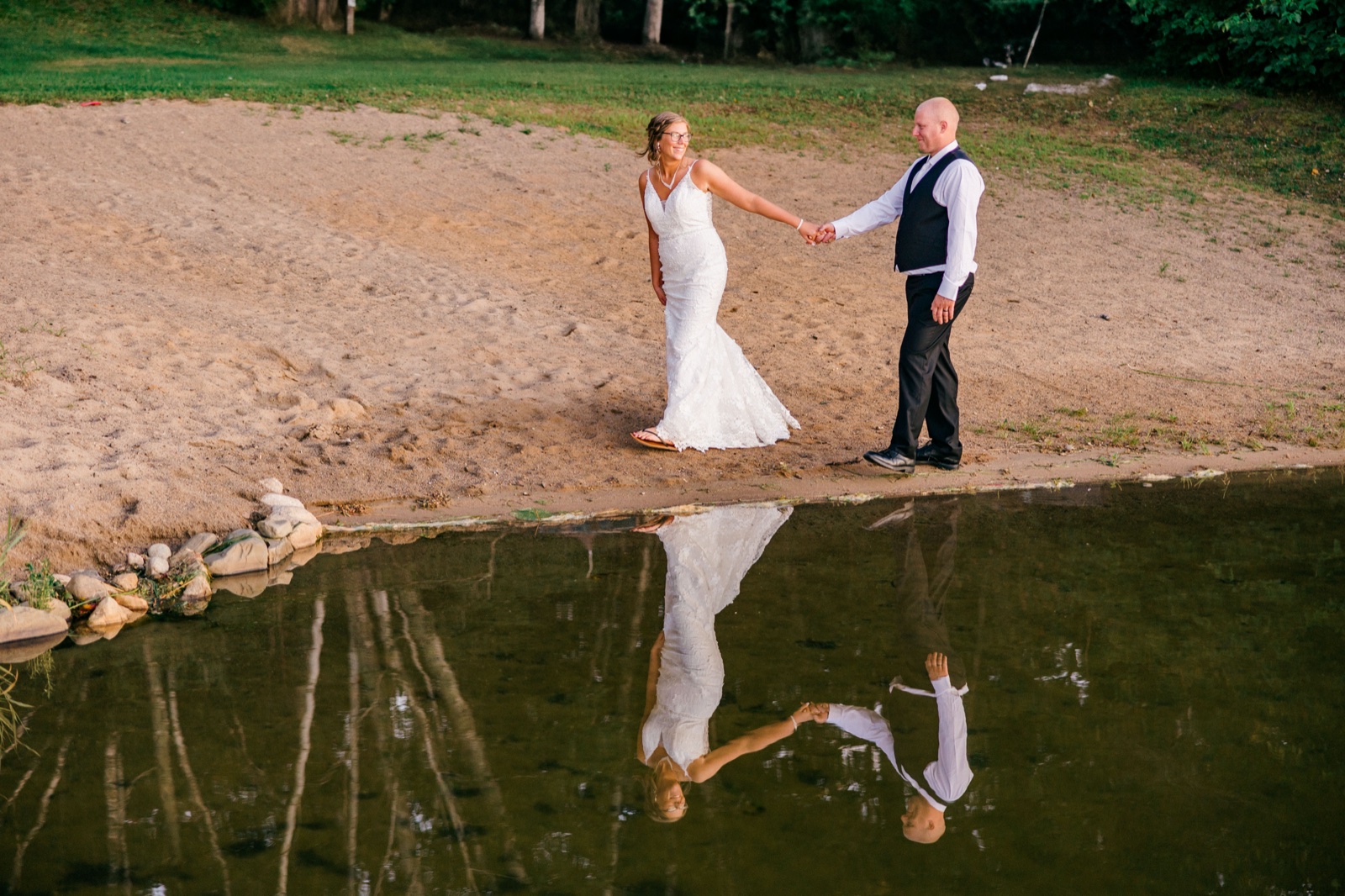 Kim and Jesse hold hands on a sandy lakeshore at golden hour, their full figures mirrored in the still water below — Tim Larsen Photography, Brainerd Lakes MN
