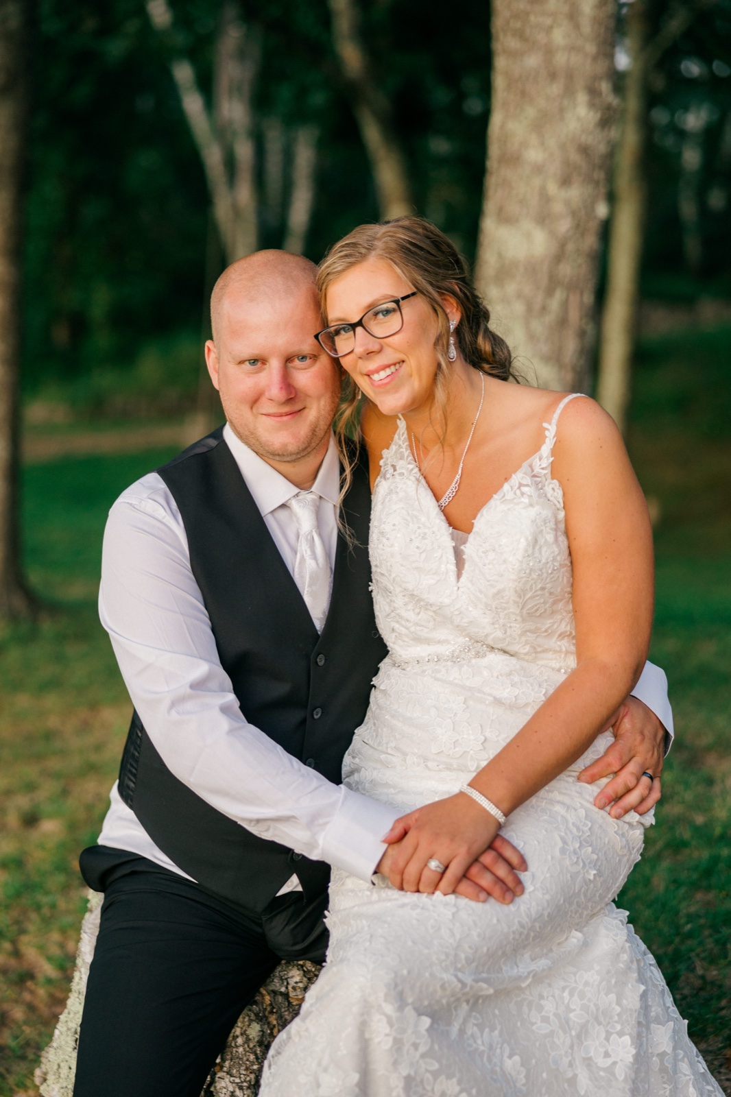Kim leans her head against Jesse as they sit together on a log in a wooded grove, both smiling at the camera in soft evening light — Tim Larsen Photography, Brainerd Lakes MN