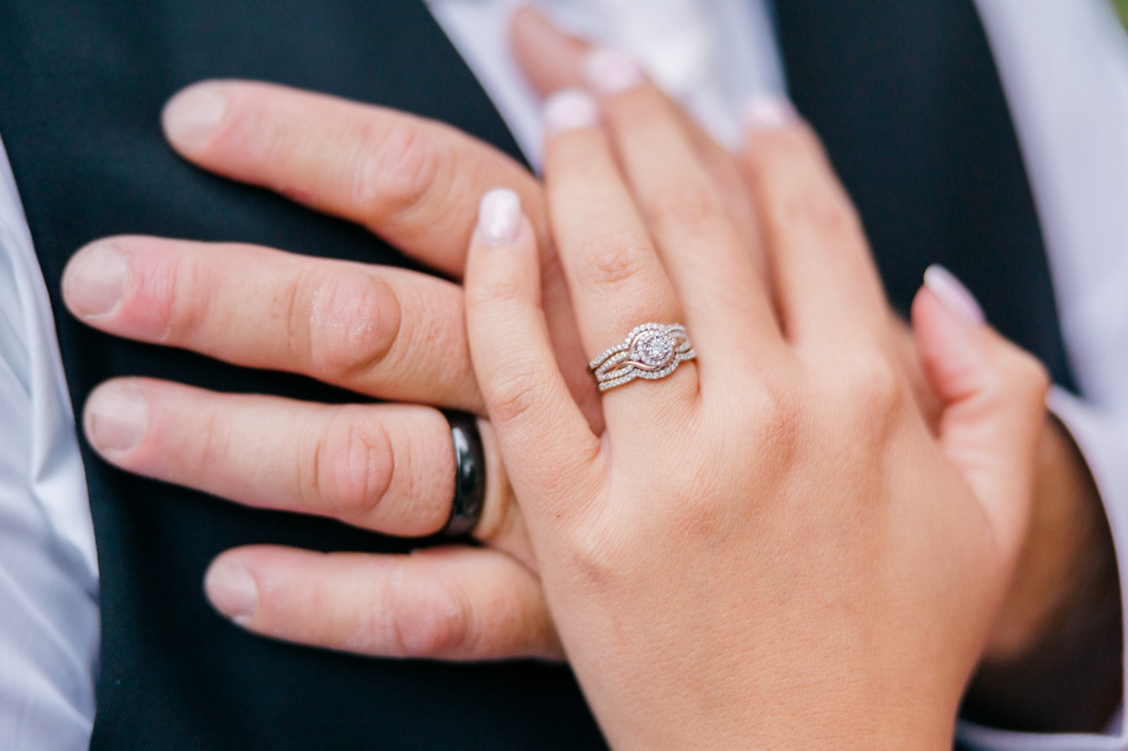 Kim's hand with her halo engagement ring and wedding band rests atop Jesse's hand, which wears a dark band, against his suit jacket — Tim Larsen Photography, Brainerd Lakes MN