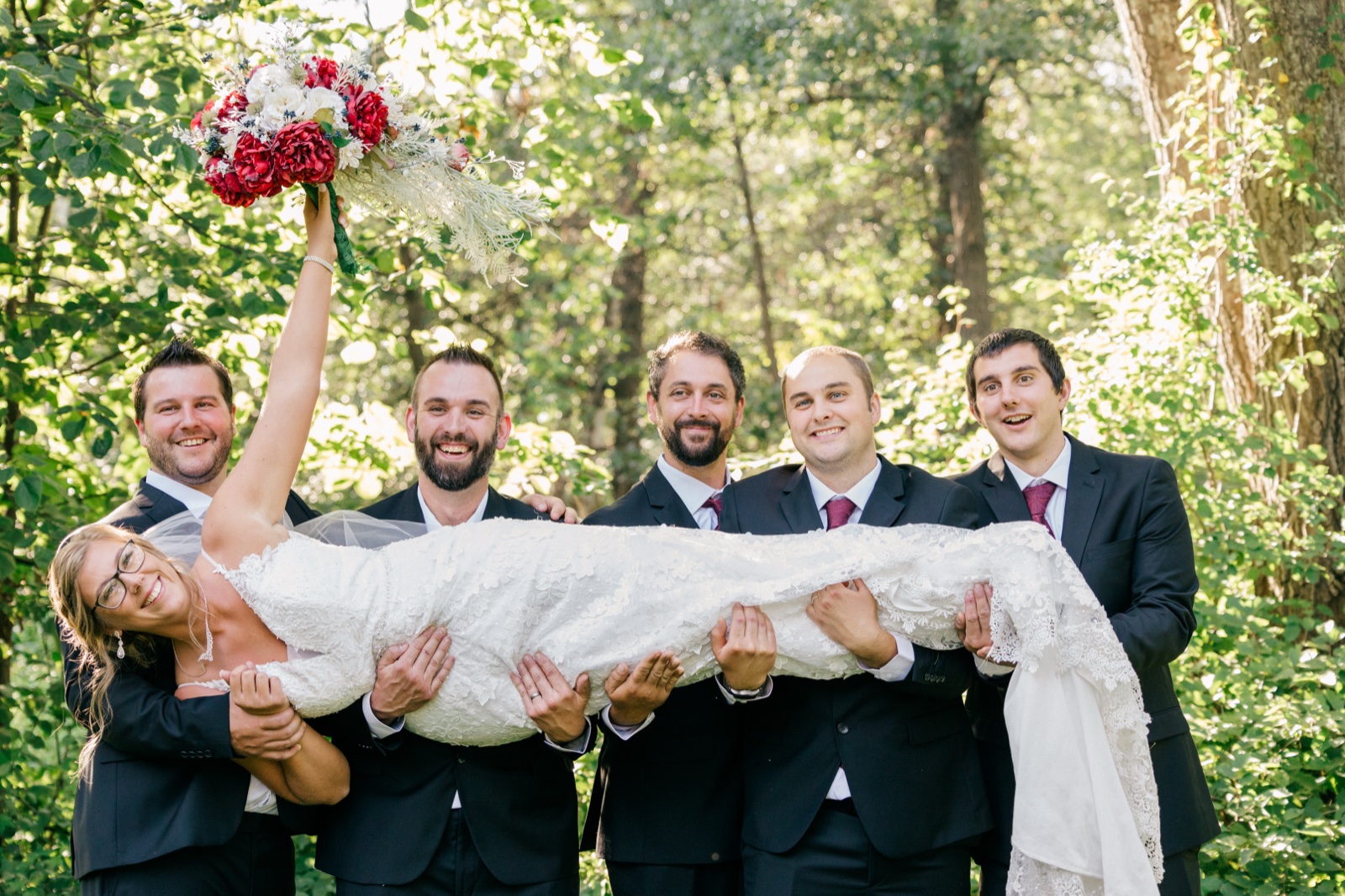 Five groomsmen in dark suits hold the bride horizontally in their arms as she raises her red and white bouquet overhead, all laughing in a sunlit forest clearing — Tim Larsen Photography, Brainerd Lakes MN
