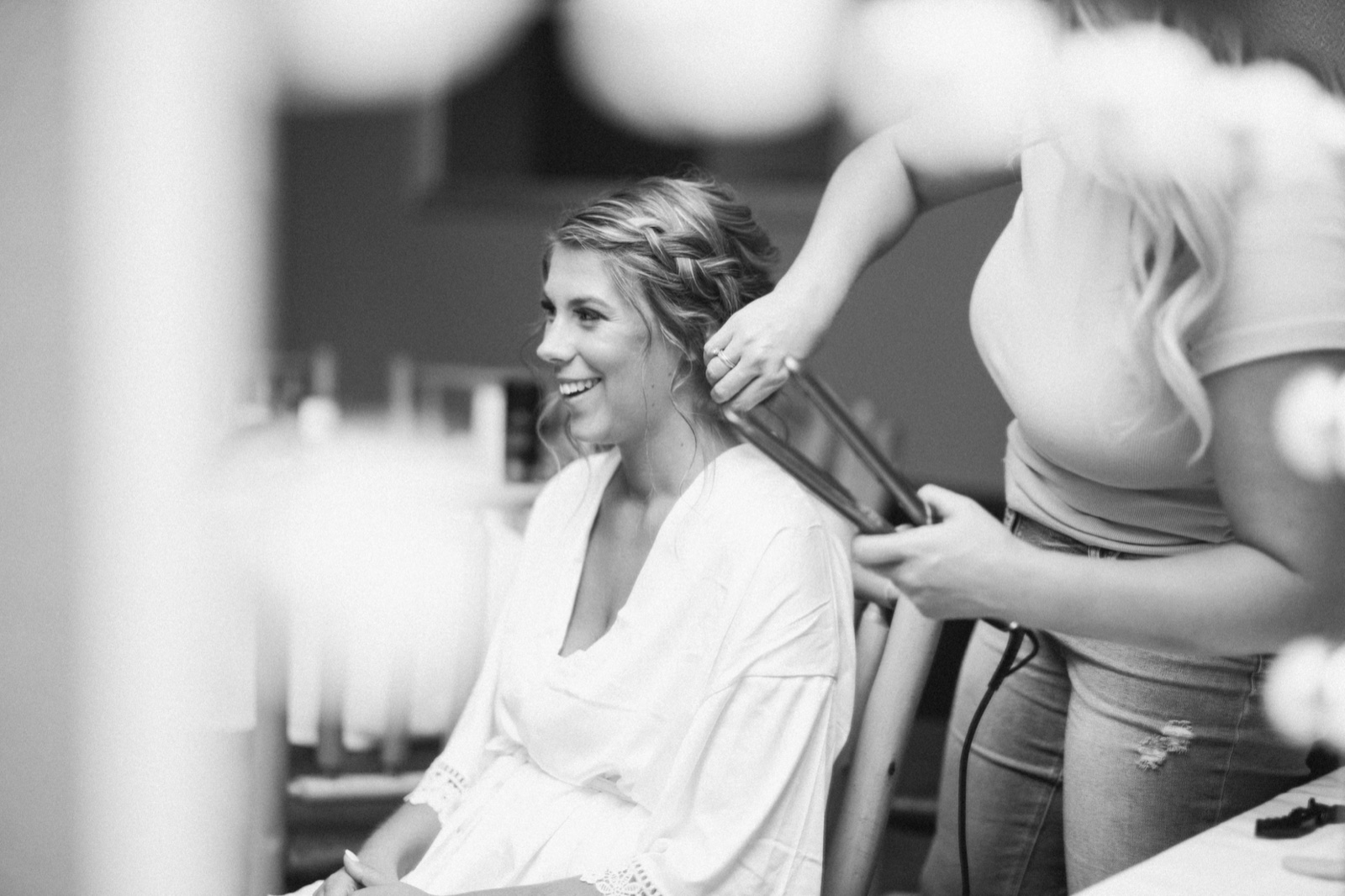 The bride laughs in a white robe while a stylist curls her hair, photographed in black and white through soft foreground bokeh — Tim Larsen Photography, Brainerd Lakes MN